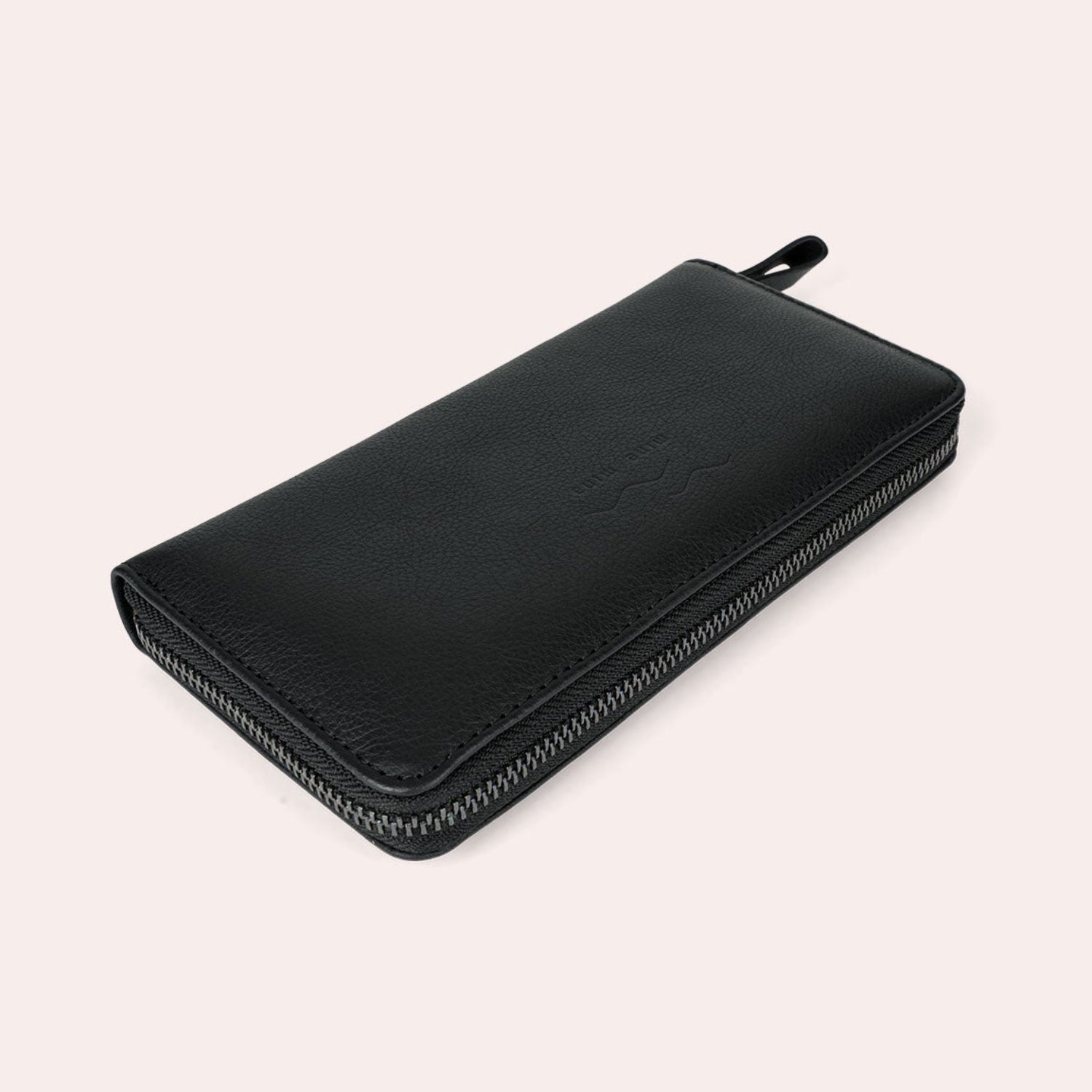 Ein Portemonnaie aus schwarzem Leder mit Reißverschluss liegt flach auf einem hellbeigen Hintergrund, dargestellt aus einer schrägen Seitenansicht. Das Portemonnaie hat ein schlichtes, minimalistisches Design mit einem sichtbaren Reißverschluss entlang der Kante.