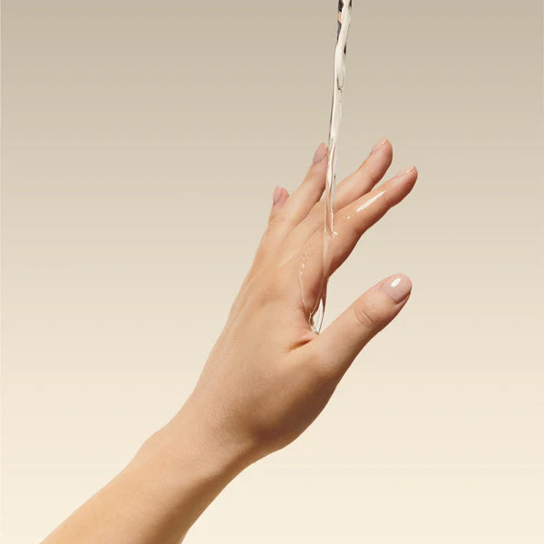 Ein Wasserstrahl fließt über den Rücken einer ausgestreckten Hand mit sauber manikürten Nägeln vor einem Hintergrund mit sanftem Beige-Verlauf.