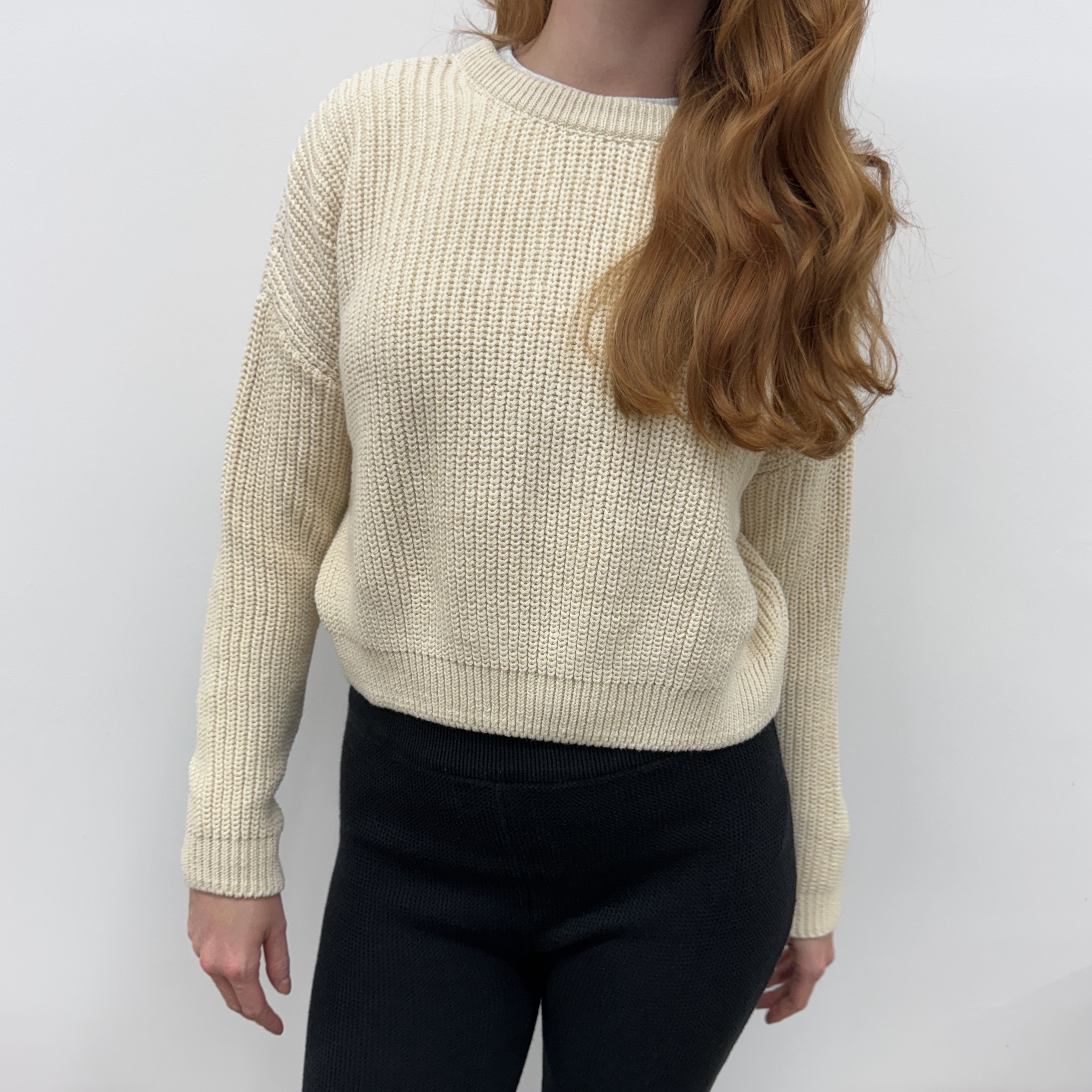 Eine Person mit langem, gewelltem, rötlich-braunem Haar trägt einen cremefarbenen Strickpullover und eine schwarze Hose und steht vor einem schlichten weißen Hintergrund. Ihr Gesicht ist teilweise außerhalb des Rahmens.