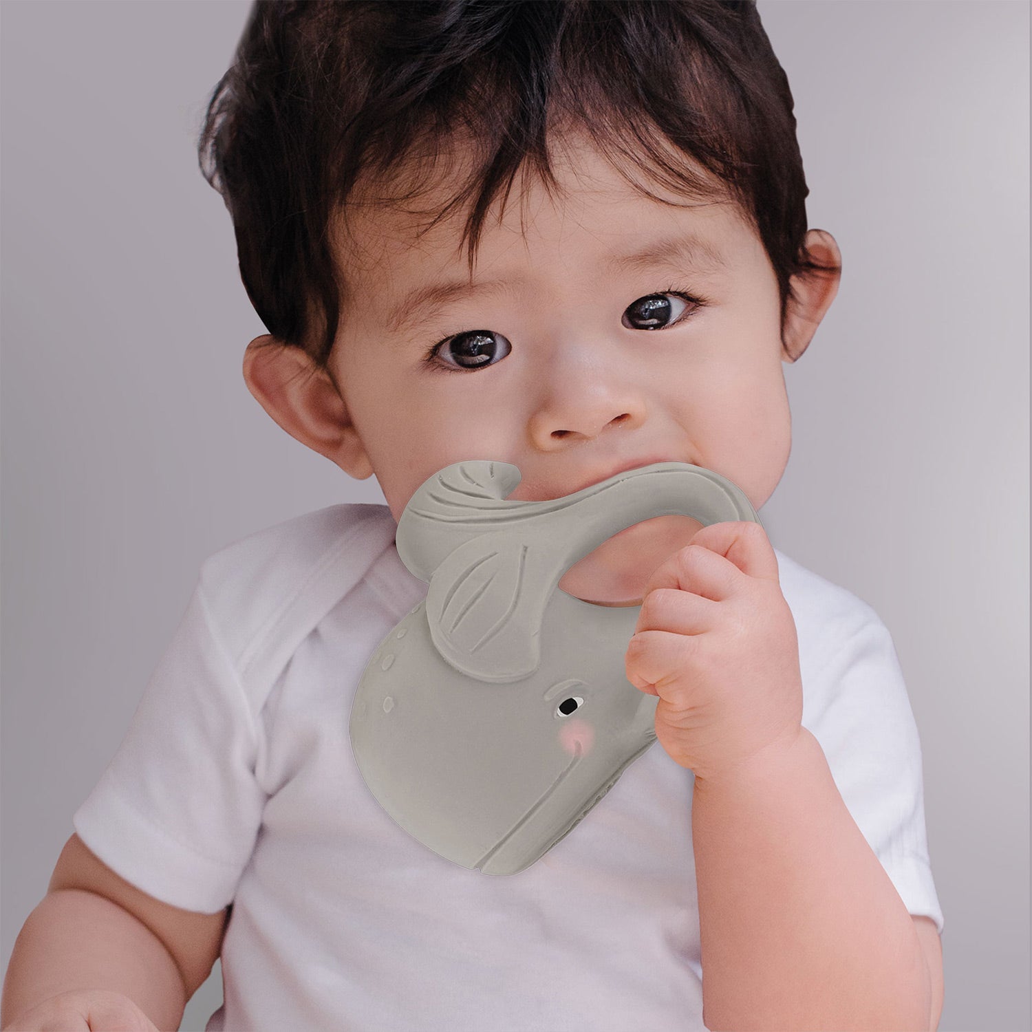 Ein Baby in einem weißen Body hält ein graues Beißspielzeug in Form eines Wals.