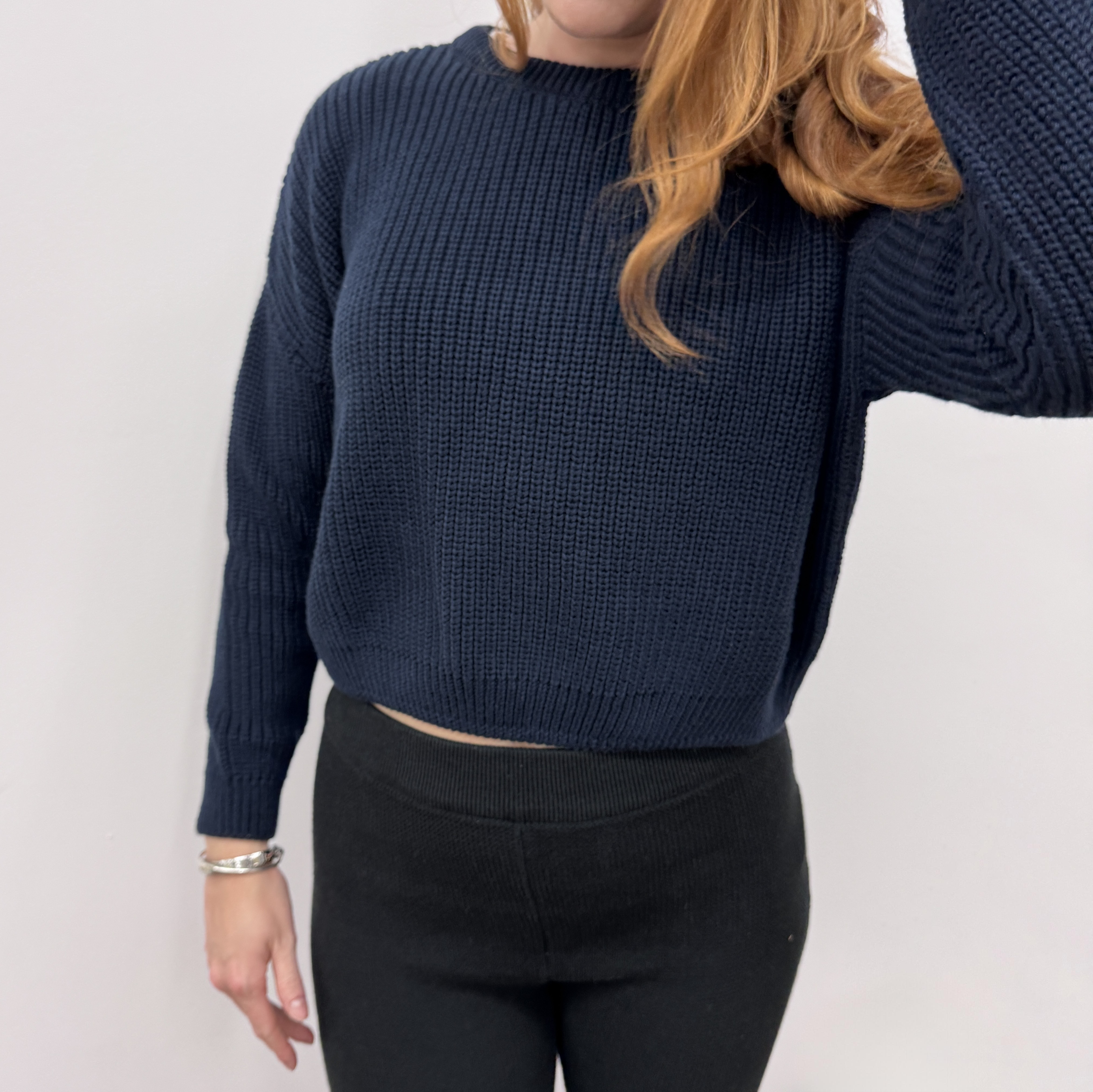 Eine Frau mit gewelltem, rötlich-blondem Haar trägt einen dunkelblauen Strickpullover und eine schwarze Hose und steht vor einem schlichten, hellen Hintergrund. Ihr Gesicht ist teilweise außerhalb des Bildausschnitts.