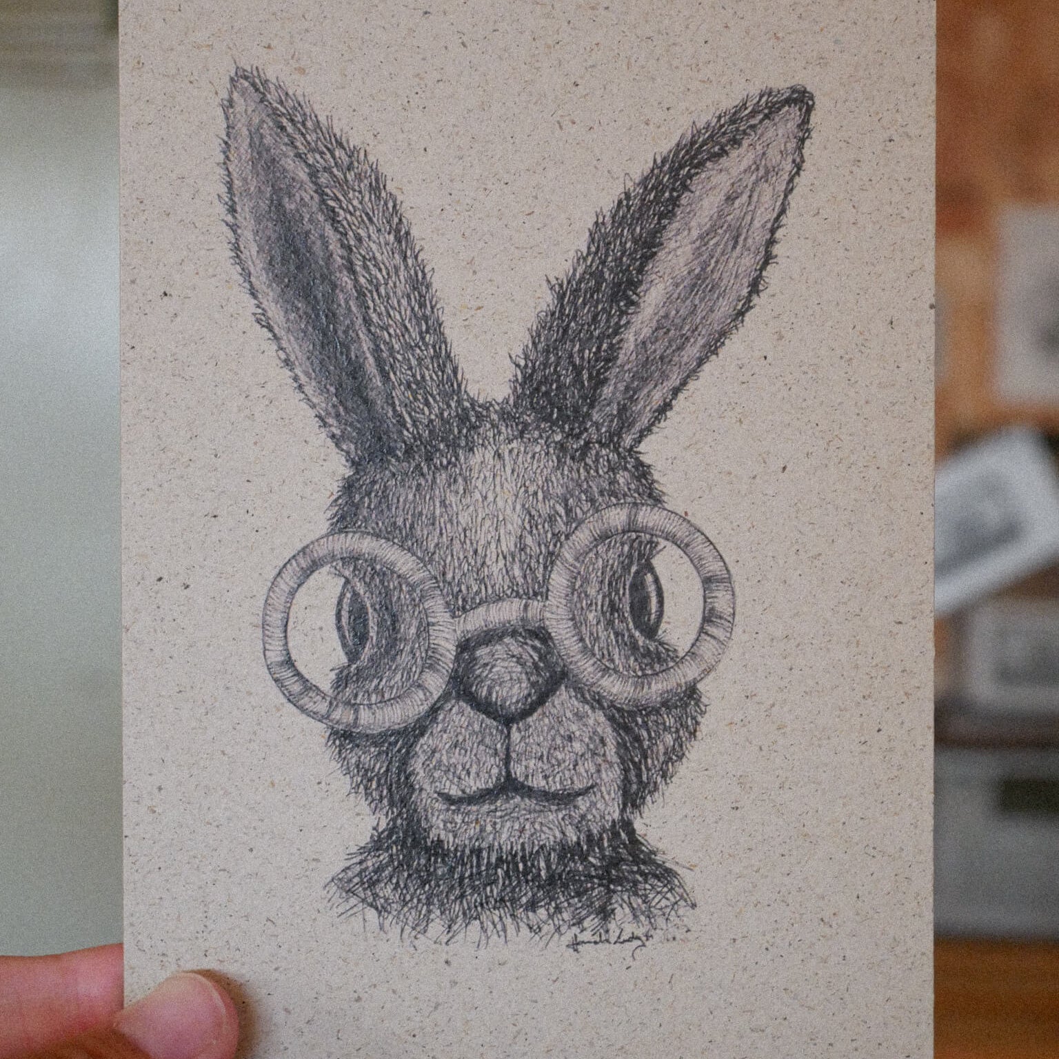 Eine Hand hält die Zeichnung eines Kaninchens mit dickem Fell und großen Ohren, das eine überdimensionale runde Brille trägt, die mit schwarzer Tinte auf beigefarbenem Strukturpapier gezeichnet ist.