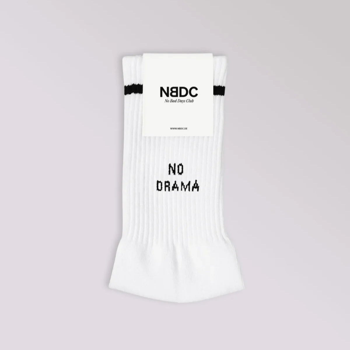 Ein Paar weiße, gerippte Socken mit schwarzen Streifen am oberen Ende und dem Text NO DRAMA auf der Vorderseite. Die Socken haben ein Etikett mit der Aufschrift NBDC No Bad Days Club auf hellgrauem Hintergrund.