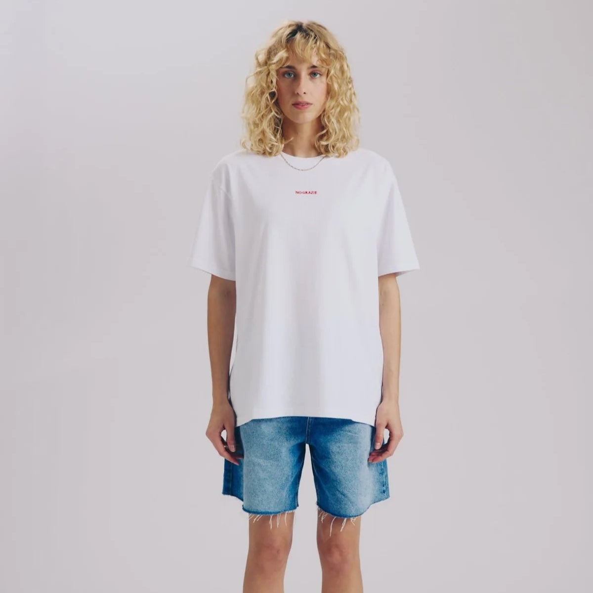 Eine Person mit lockigem blondem Haar steht mit dem Gesicht nach vorne und trägt ein lockeres weißes T-Shirt, blaue Jeansshorts, weiße Socken und weiße Turnschuhe vor einem schlichten hellen Hintergrund.