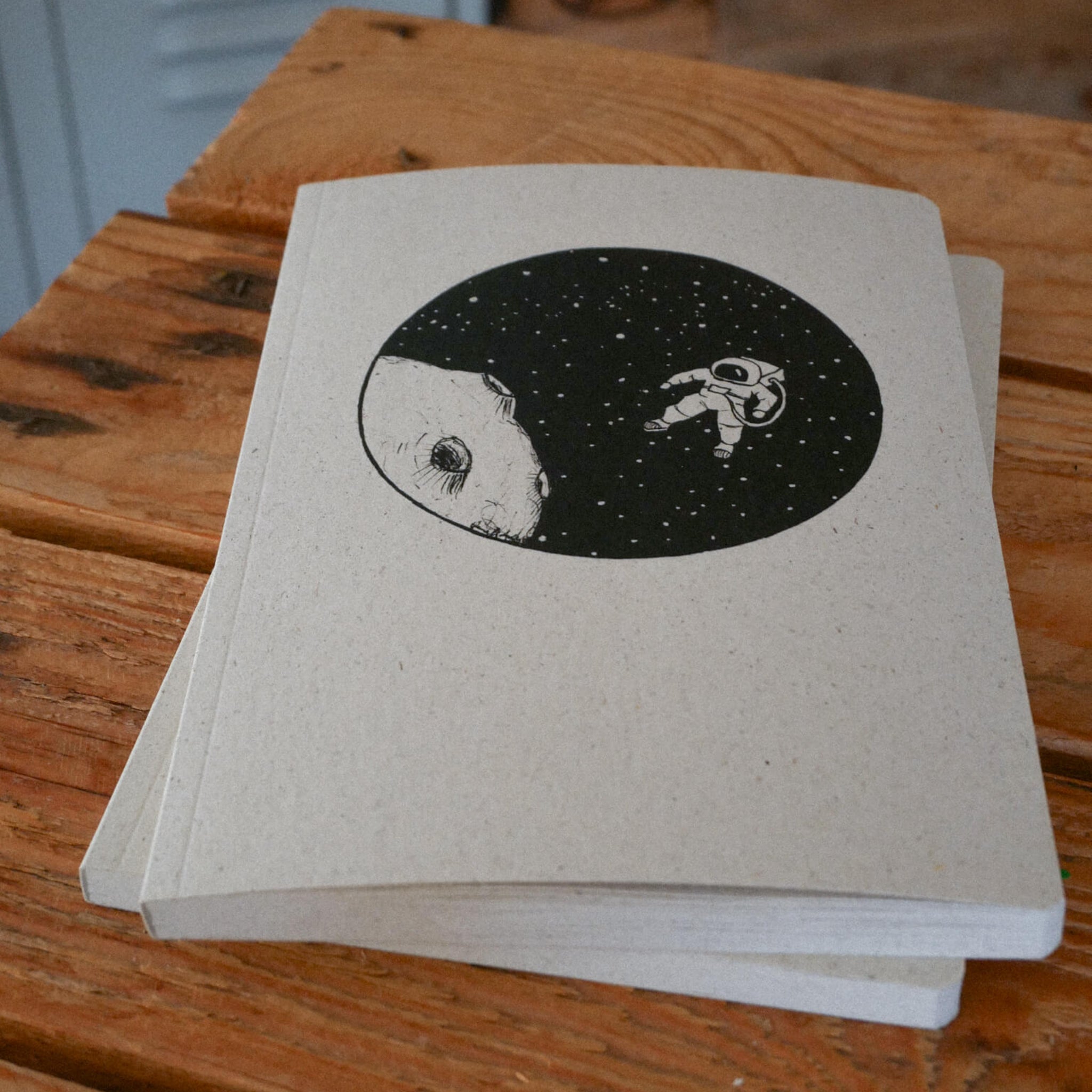 Ein Notizbuch mit der Illustration eines im Weltraum schwebenden Astronauten in der Nähe eines großen Tiergesichts vor einem schwarzen kreisförmigen Hintergrund liegt auf einer Holzfläche.