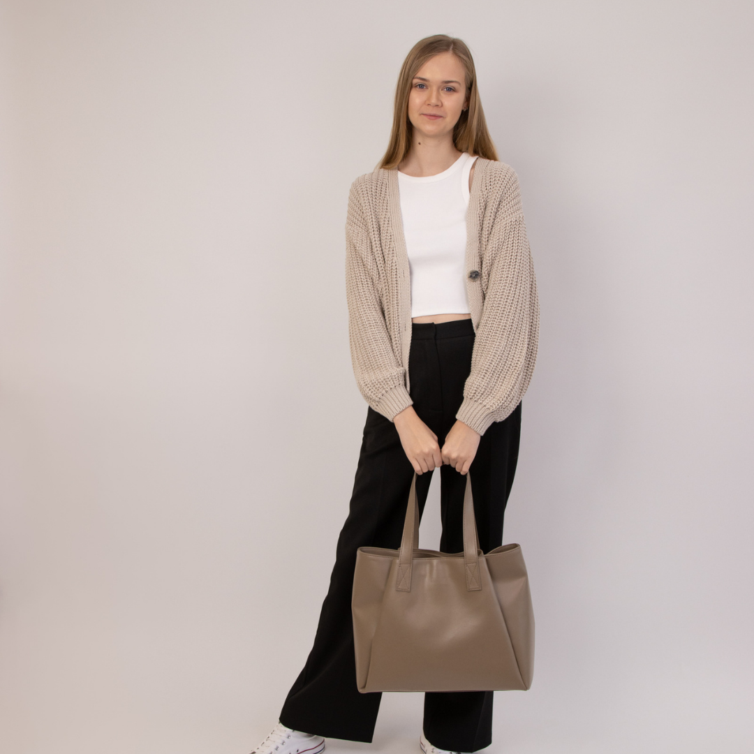 Frau in beigem Cardigan und schwarzer Hose hält eine braune Tasche vor einem neutralen Hintergrund.