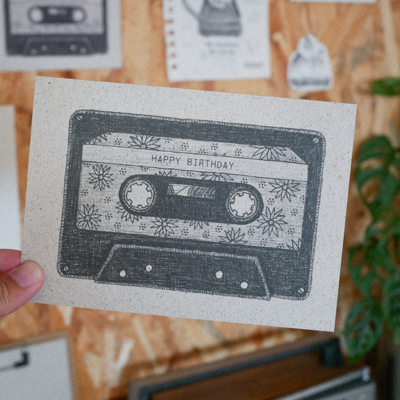 Eine Hand hält eine Karte mit einer Schwarz-Weiß-Zeichnung einer Kassette mit der Aufschrift Happy Birthday und dekorativen Sternen. Im Hintergrund sind weitere illustrierte Karten und eine Grünpflanze zu sehen.