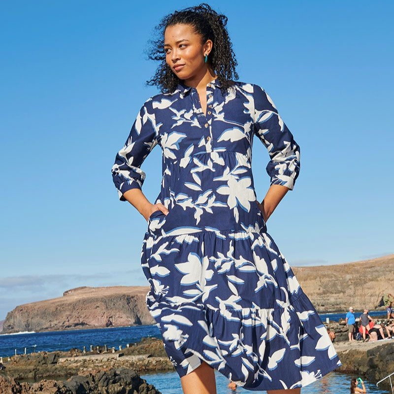 Frau in blau-weiß gemustertem Kleid steht am Meer vor Felsen und klarem Himmel.