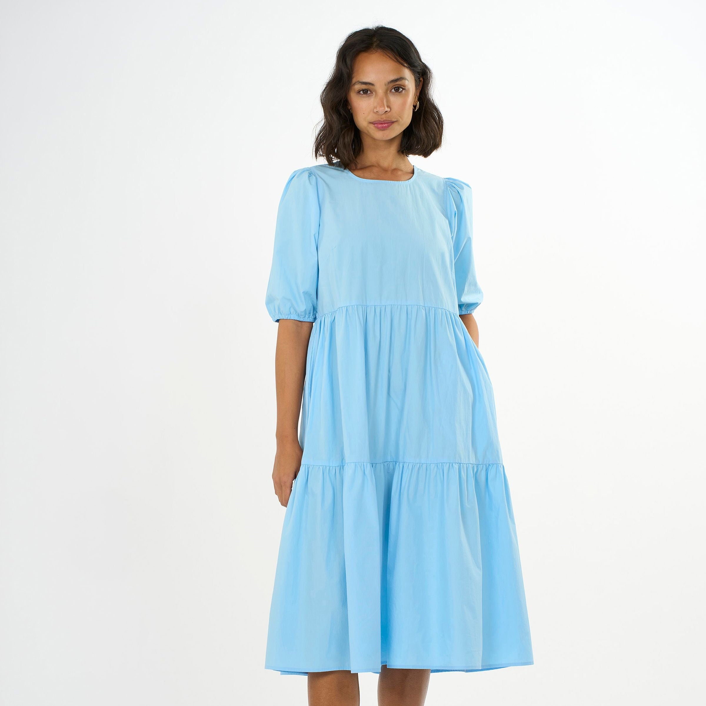 Frau in einem hellblauen, locker sitzenden Kleid mit halblangen Ärmeln und Rüschen vor weißem Hintergrund.
