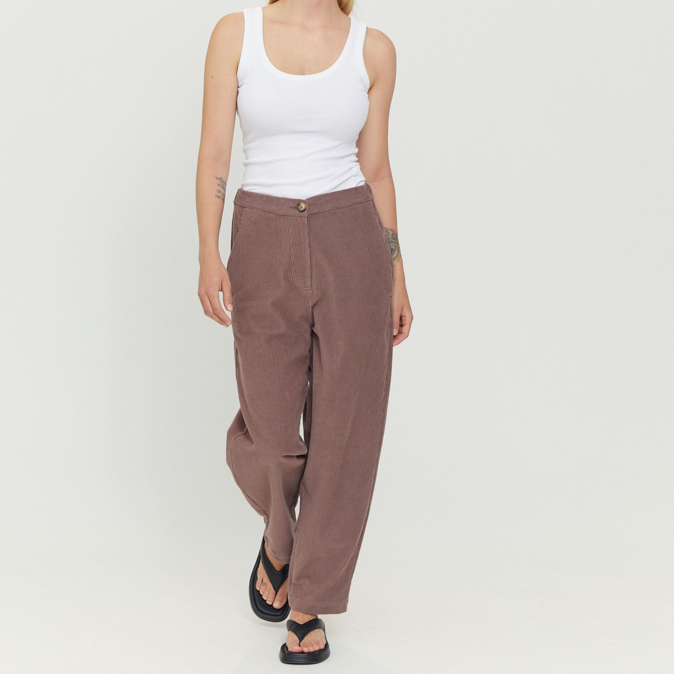 Frau in weißem Tanktop und brauner Hose, die Sandalen trägt, geht auf hellem Hintergrund.