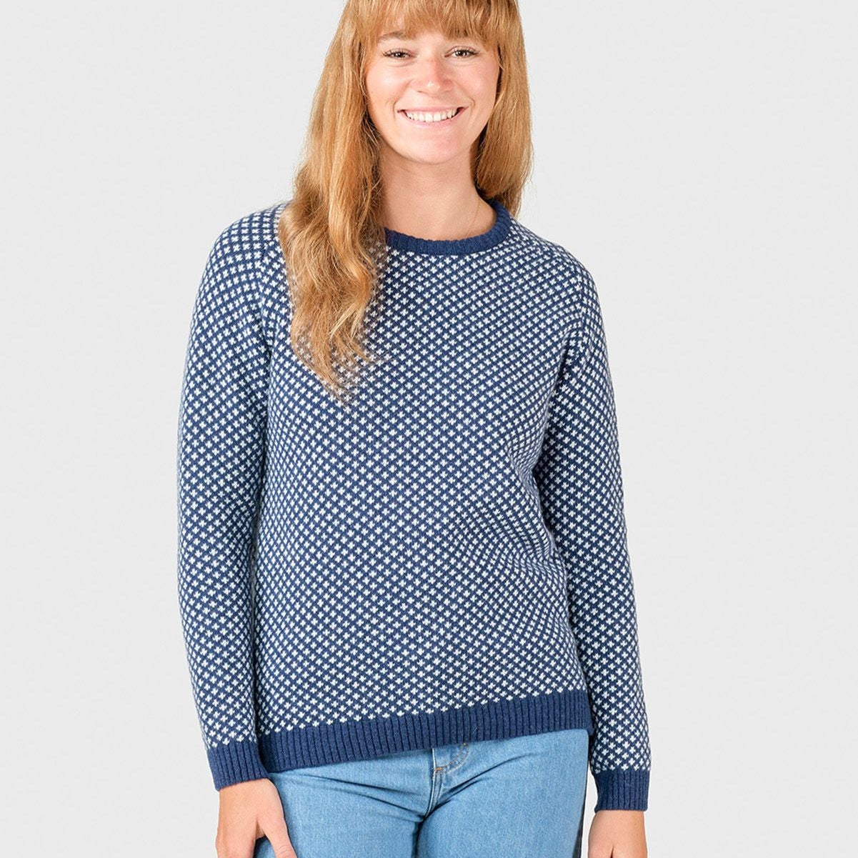 Lächelnde Person in blau-weiß gemustertem Pullover und blauer Jeans vor neutralem Hintergrund.