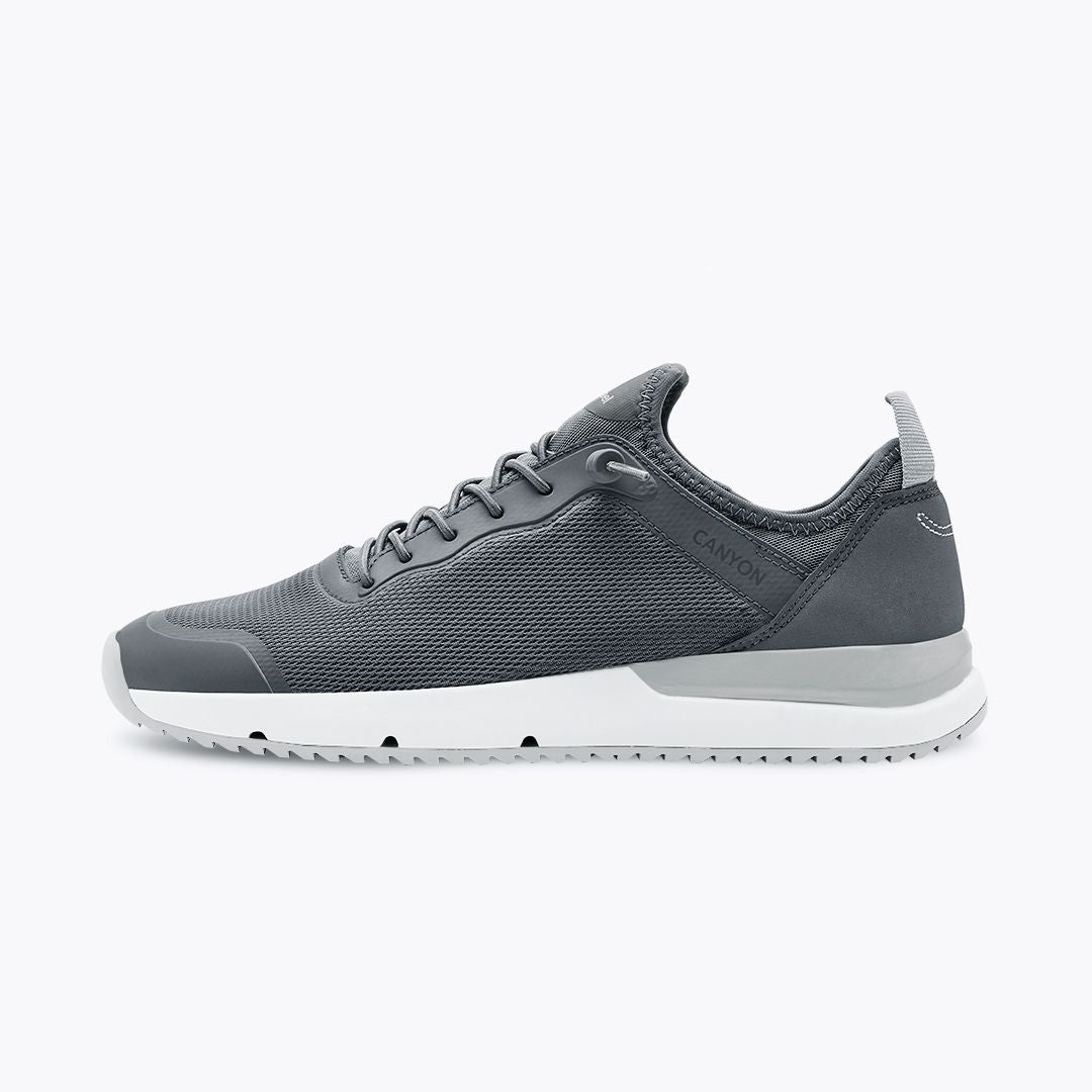 Dunkelgrauer Sneaker mit weißer Sohle auf weißem Hintergrund, seitlich abgebildet, mit Netzdetails und Schnürsenkeln.