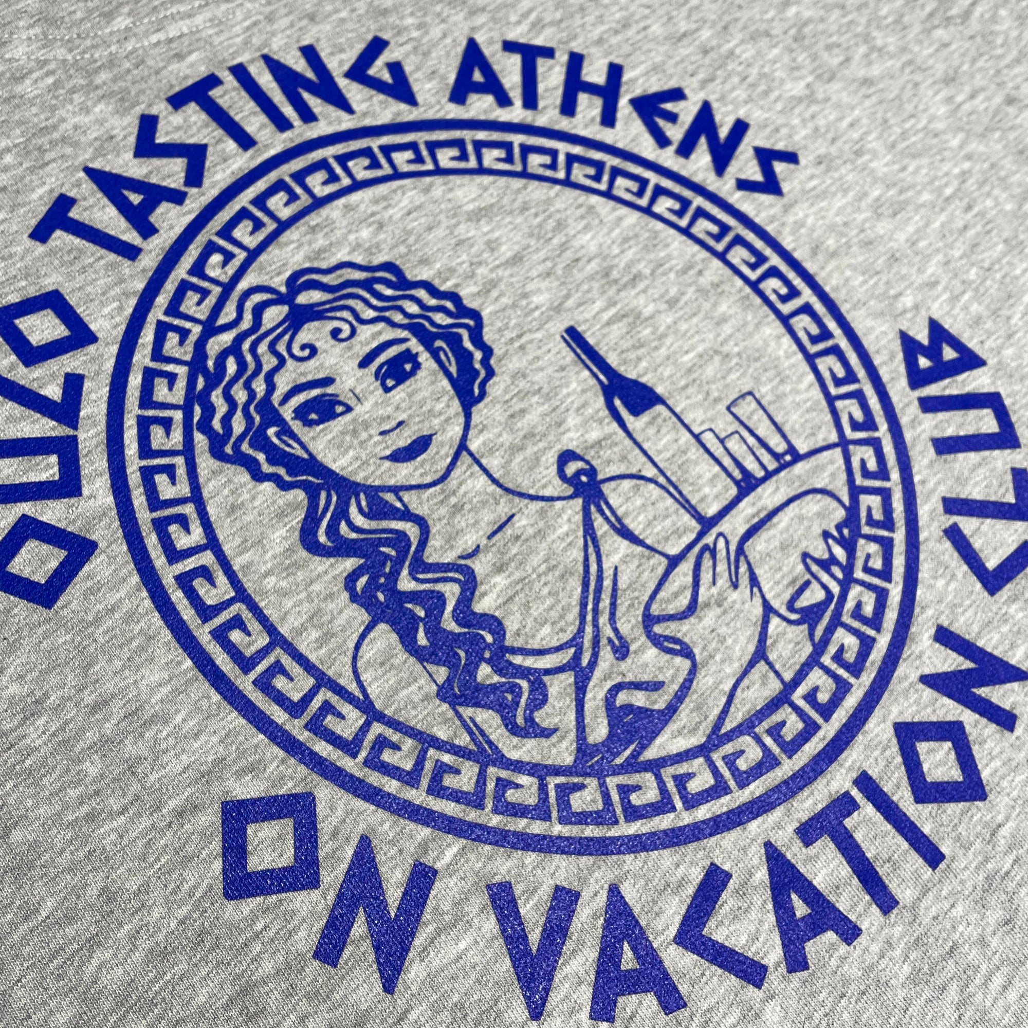 Griechisches Design auf einem grauen T-Shirt, zeigt eine Frau mit einer Flasche Ouzo und Brot, umgeben von geometrischem Muster.