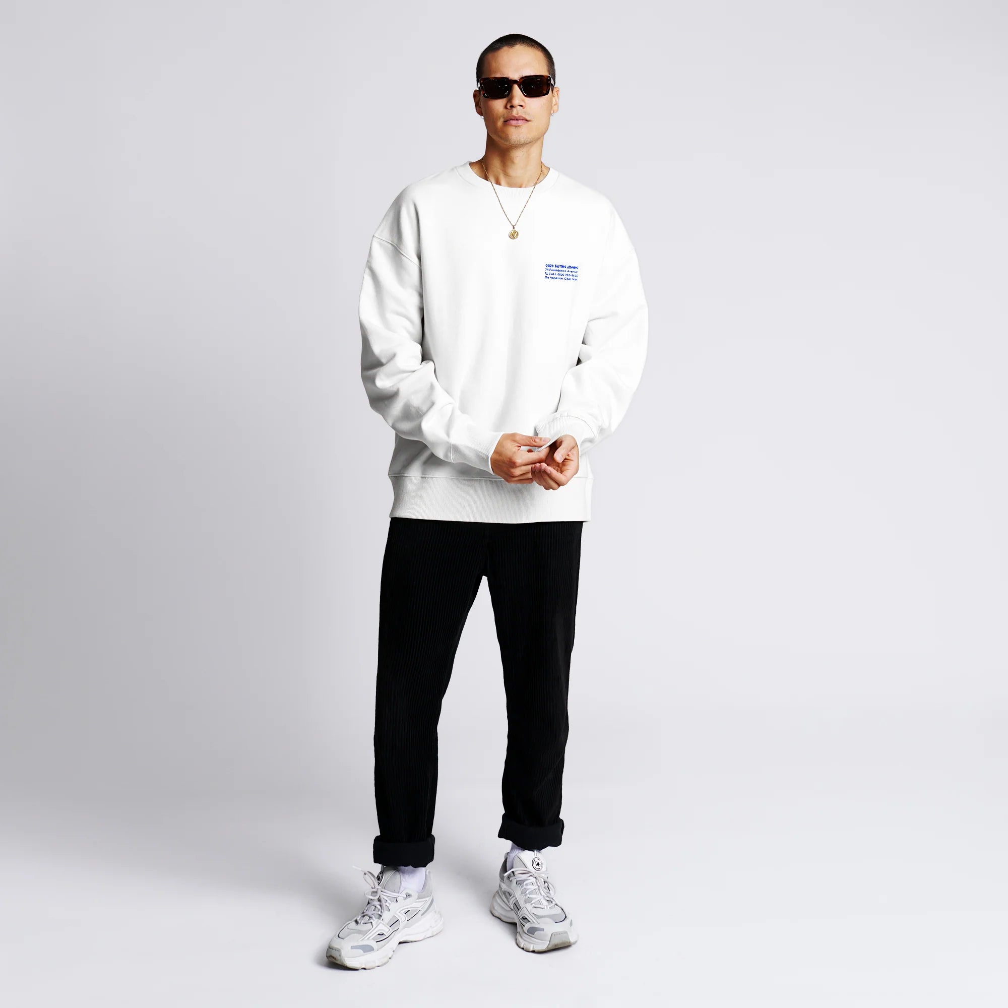 Person in weißem Pullover, schwarzer Hose und Sneakers steht vor weißem Hintergrund.