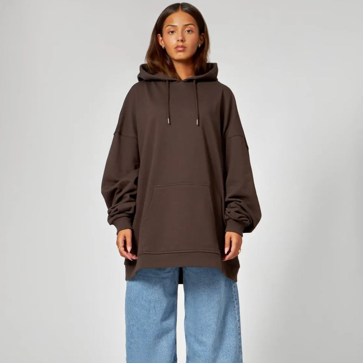 Eine Person steht mit dem Gesicht nach vorne, trägt einen dunkelbraunen, übergroßen Kapuzenpullover, hellblaue, weit geschnittene Jeans und schwarze Sandalen vor einem schlichten grauen Hintergrund.