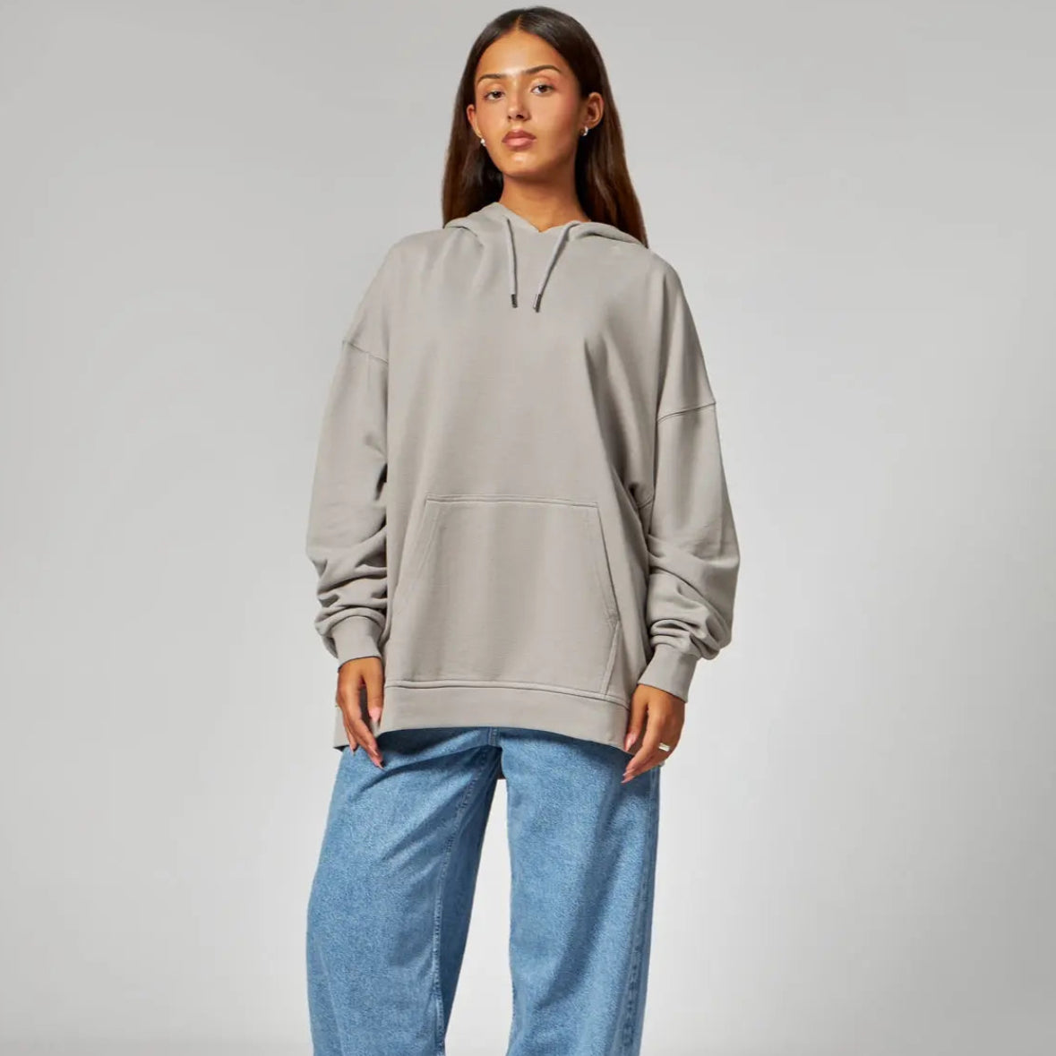 Eine Frau steht vor einem schlichten Hintergrund und trägt einen lockeren grauen Kapuzenpulli, weite Bluejeans und schwarze Schuhe. Sie hat langes braunes Haar und einen neutralen Gesichtsausdruck.