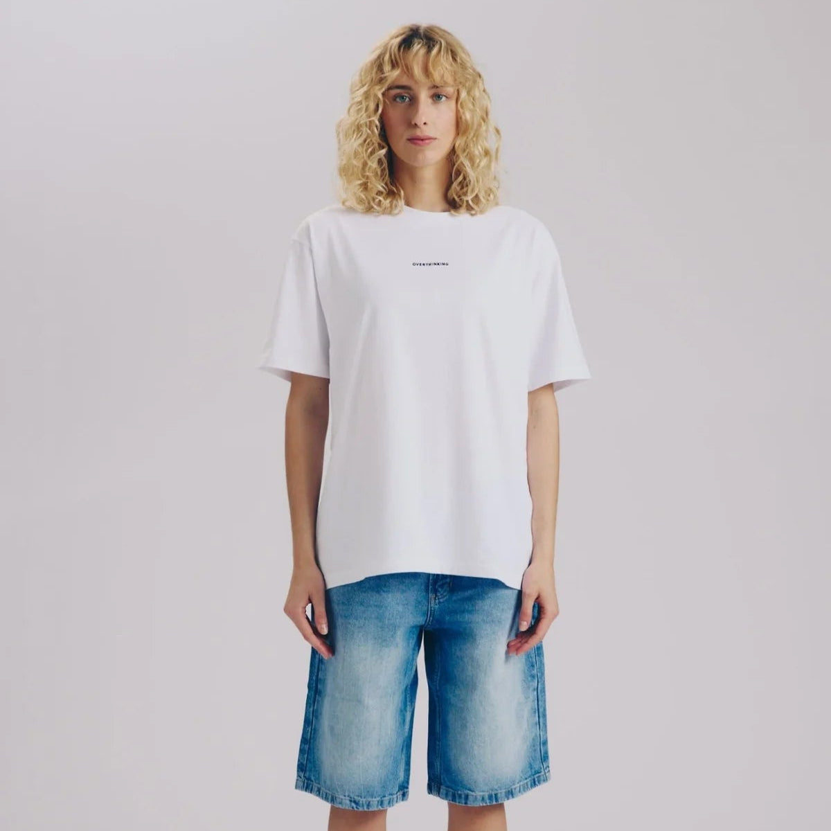 Eine Person mit lockigem blondem Haar steht mit dem Gesicht nach vorne und trägt ein lockeres weißes T-Shirt, knielange Jeans-Shorts, weiße Socken und weiße Turnschuhe vor einem schlichten, hellen Hintergrund.