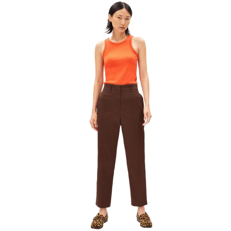 Eine Frau steht vor einem weißen Hintergrund und trägt ein ärmelloses orangefarbenes Oberteil, eine hochtaillierte braune Hose und Slipper mit Leopardenmuster. Sie hat glattes schulterlanges dunkles Haar und einen neutralen Gesichtsausdruck.