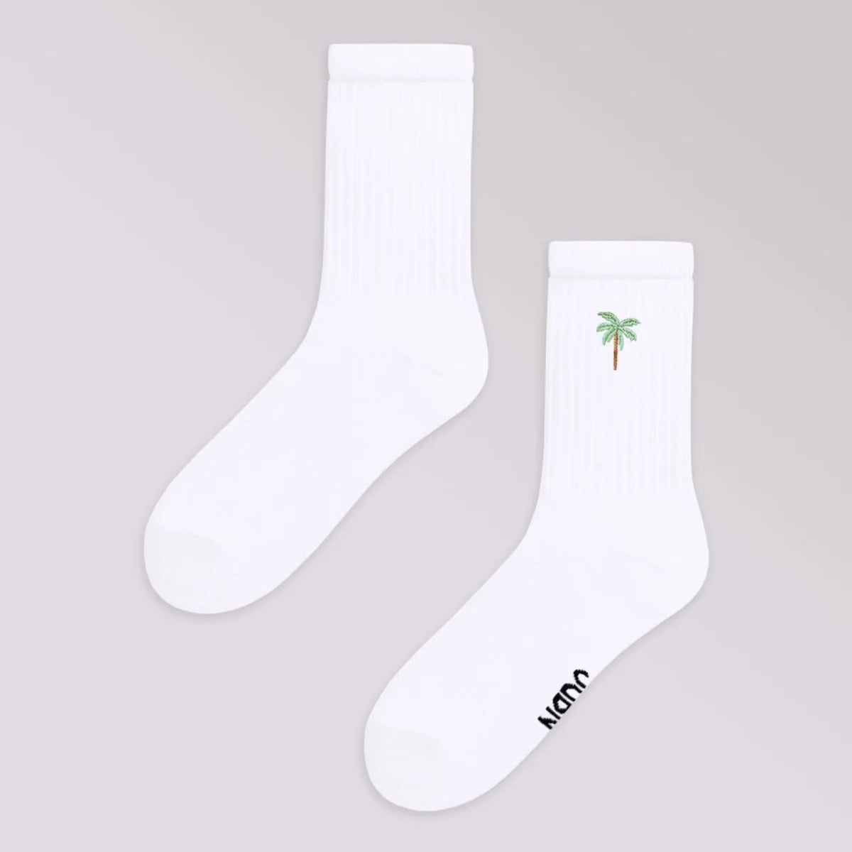 Ein Paar weiße Socken auf einem hellgrauen Hintergrund. Eine Socke zeigt eine kleine gestickte grüne Palme in der Nähe der Spitze, während die andere Socke hat schwarzen Text teilweise auf der Sohle sichtbar.