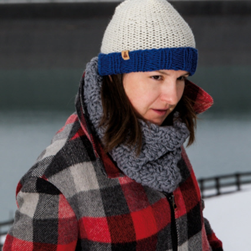 Eine Frau mit einer weiß-blauen Strickmütze, einem grauen Schal und einem rot-schwarz karierten Mantel steht im Freien in einer verschneiten Umgebung am Wasser.