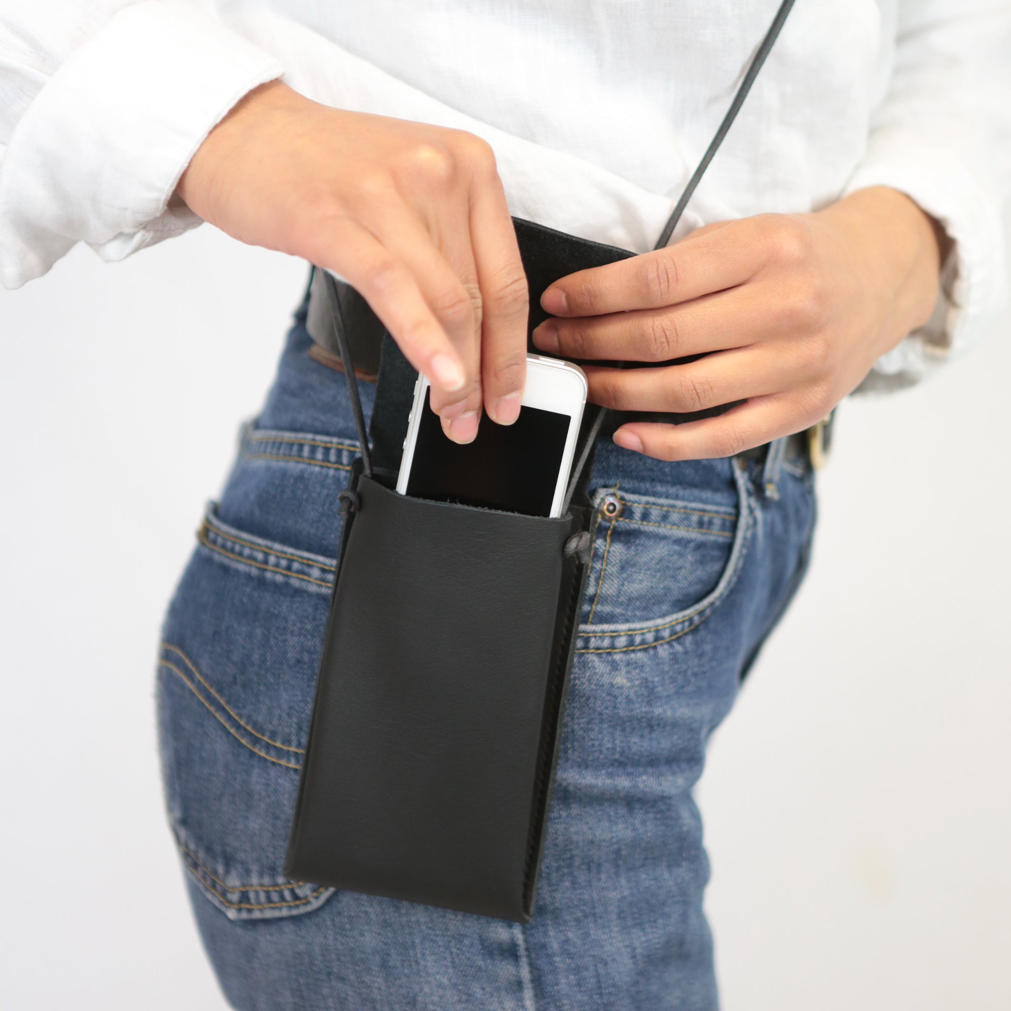 Person steckt ein Smartphone in eine schwarze Lederhülle, die an einem Gürtel befestigt ist, während sie Jeans trägt.