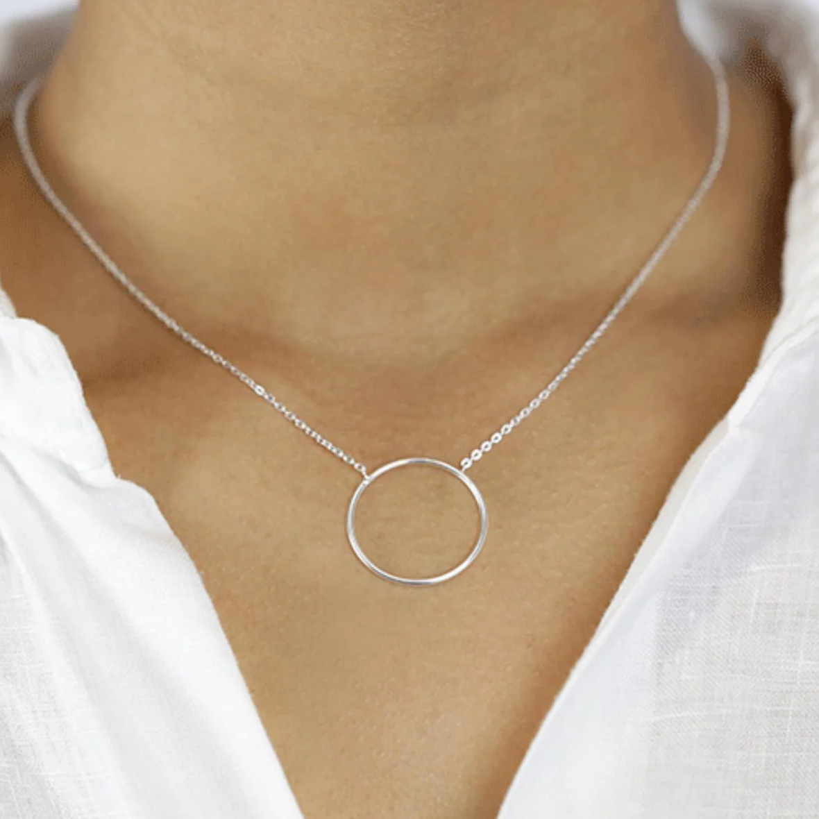 Silberne Halskette mit rundem Anhänger auf nacktem Hals, getragen über einer weißen Bluse.