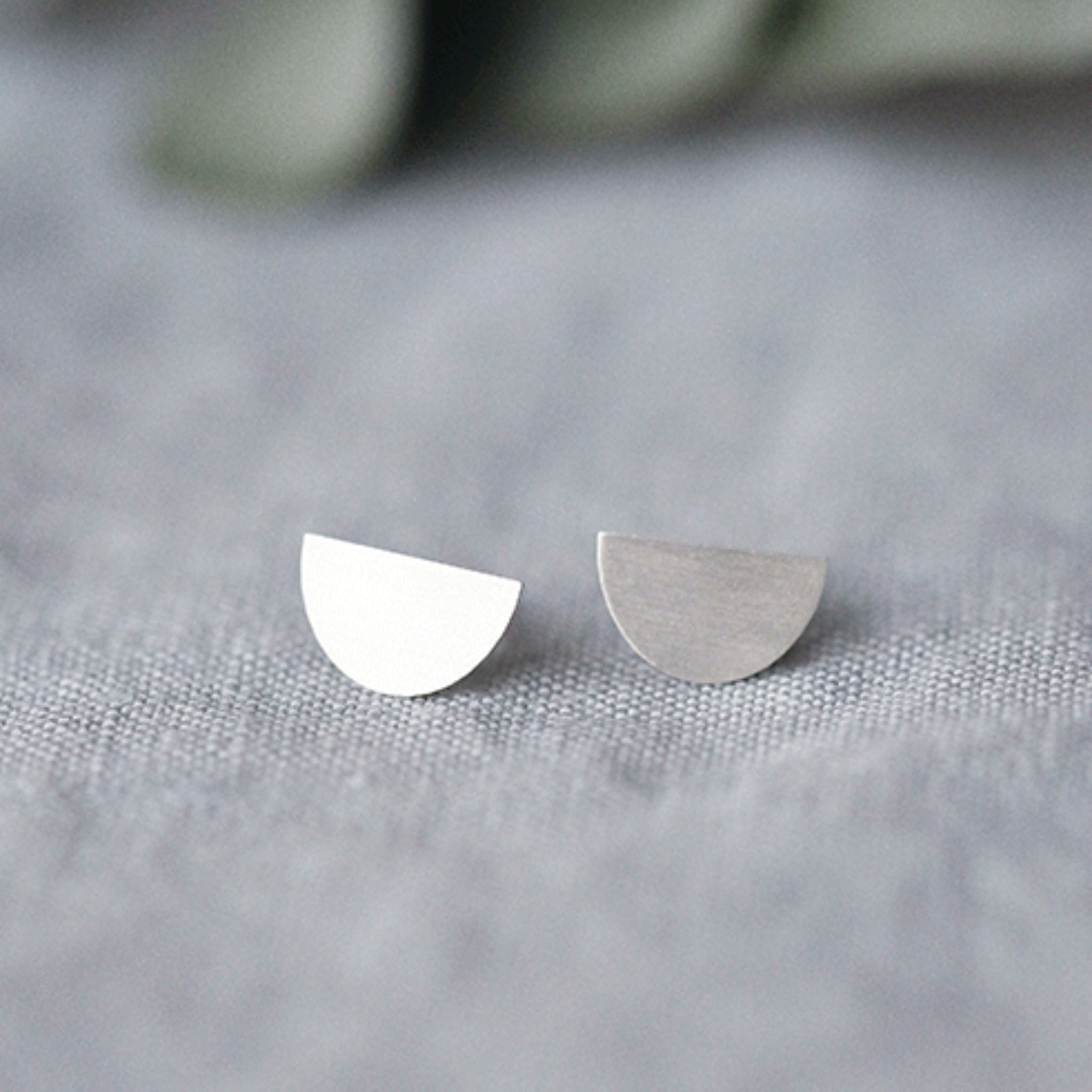 Zwei minimalistische halbkreisförmige Ohrstecker, einer in glänzendem Silber, der andere in mattem Silber, auf einem Hintergrund aus strukturiertem grauem Stoff.