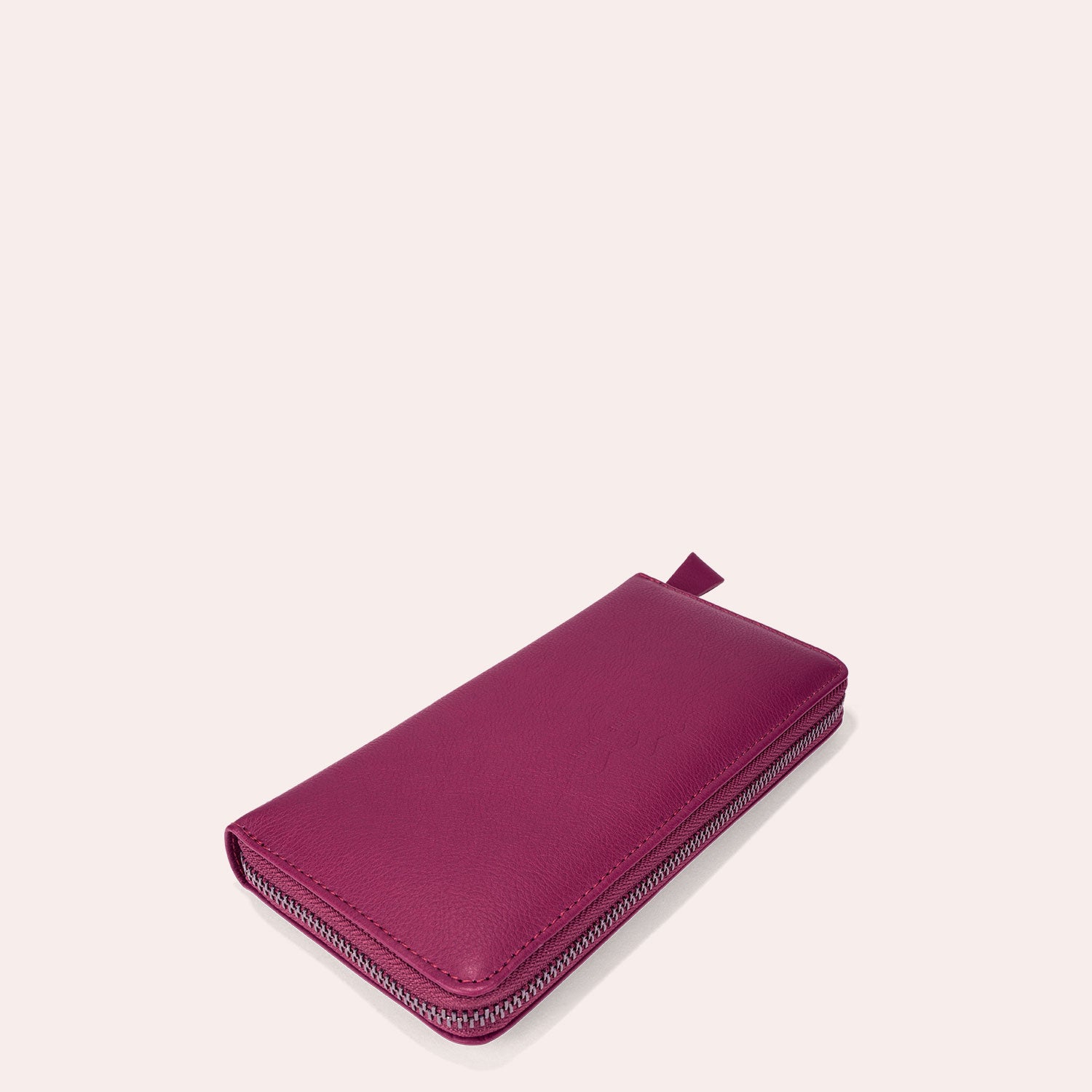 Ein magentafarbenes Lederportemonnaie mit Reißverschluss und Zuglasche, das geschlossen auf einem hellrosa Hintergrund dargestellt wird.