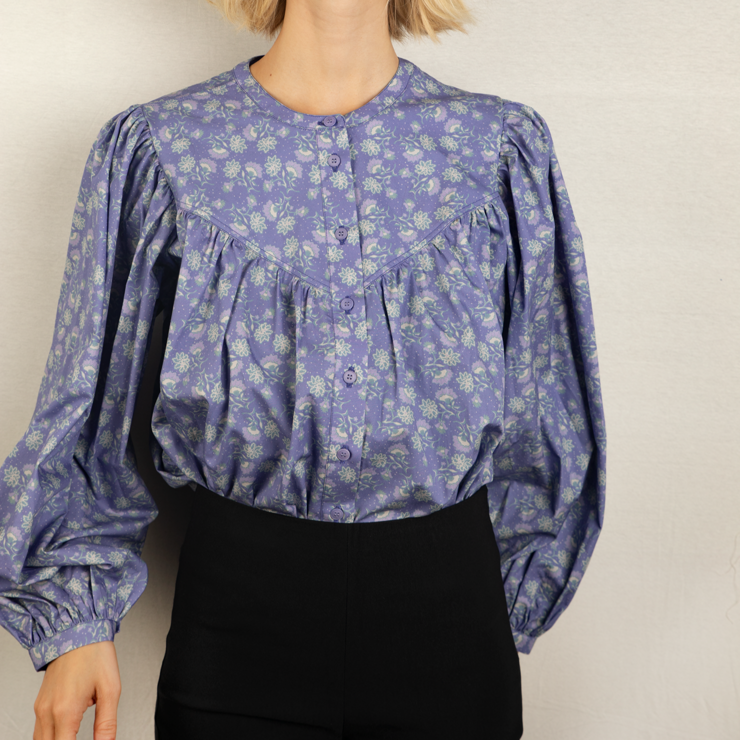 Frau in lila gemusterter Bluse mit langen Puffärmeln und schwarzer Hose vor neutralem Hintergrund.