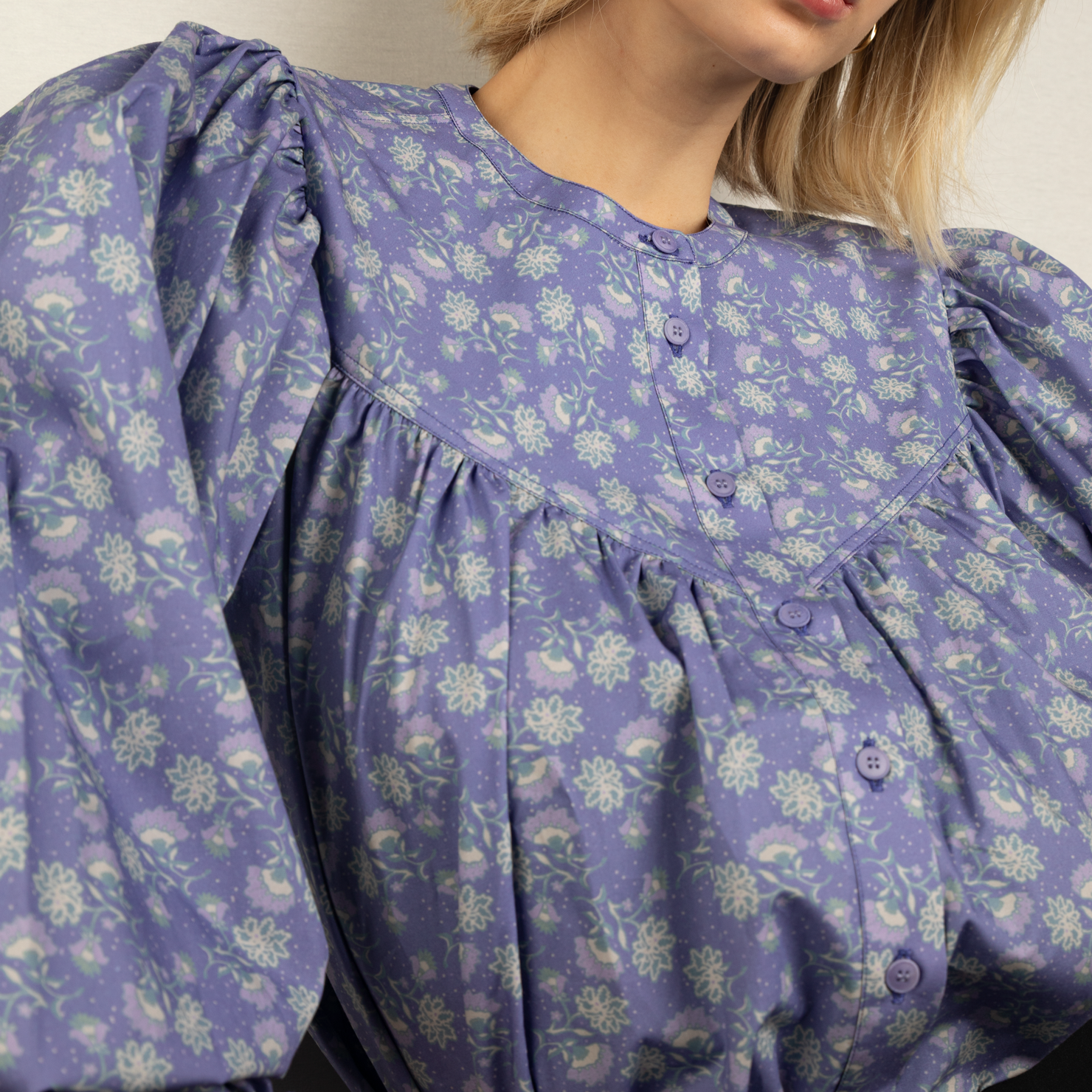 Eine Person in einer lila Bluse mit floralem Muster und Puffärmeln, im Porträt aufgenommen.