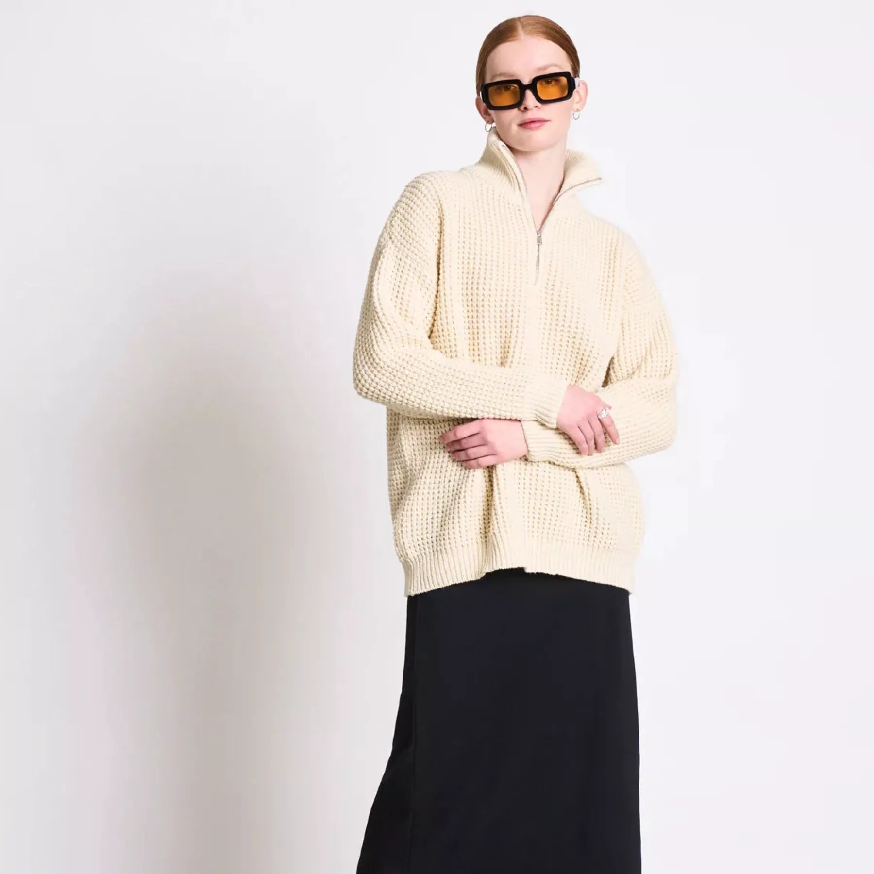 Eine Person, die einen dicken cremefarbenen Pullover mit Reißverschluss, einen langen schwarzen Rock, schwarze Stiefel und eine übergroße Sonnenbrille trägt, steht mit locker verschränkten Händen vor einer schlichten weißen Wand.