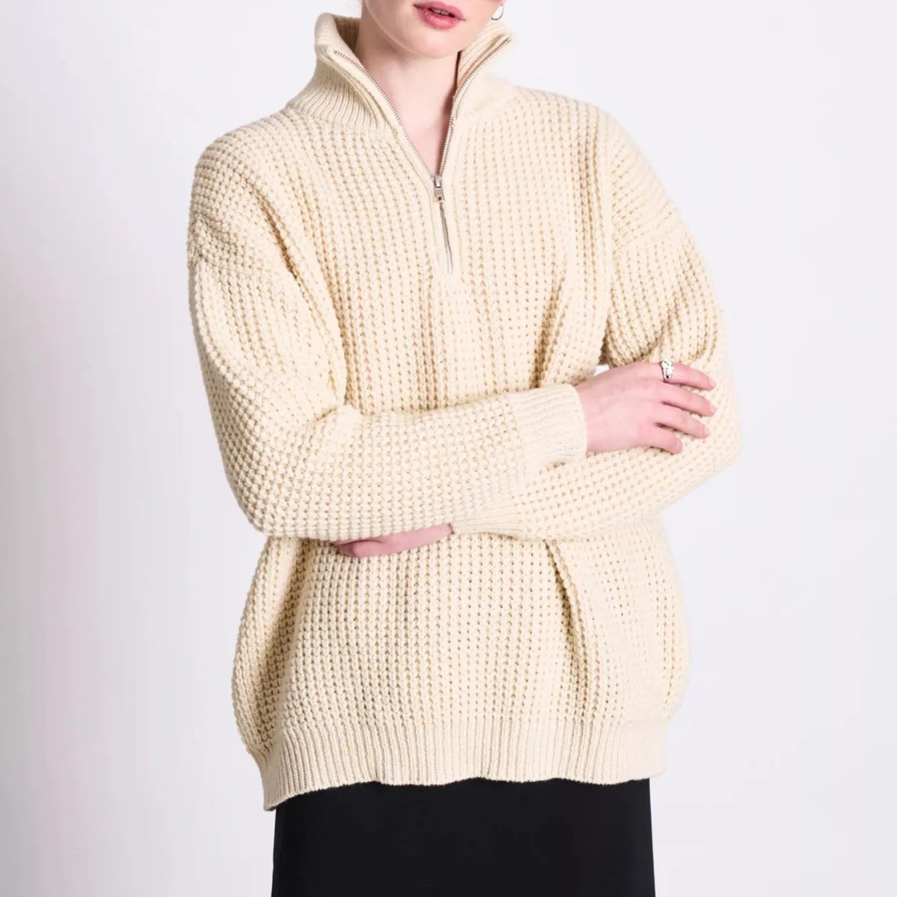 Eine Frau mit rotem Haar in einem niedrigen Pferdeschwanz trägt eine rechteckige Sonnenbrille, Perlenohrringe, einen cremefarbenen Strickpullover mit Reißverschluss und einen schwarzen Rock. Sie steht mit verschränkten Armen vor einem einfarbigen Hintergrund.