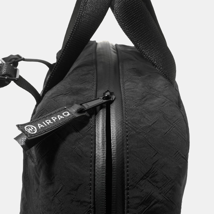 Nahaufnahme einer schwarzen Tasche mit strukturierter Oberfläche, die einen teilweise geöffneten Reißverschluss und ein Etikett mit der Aufschrift AIRPAQ aufweist, das am Reißverschluss befestigt ist. An der Oberseite sind stabile Tragegriffe zu sehen.
