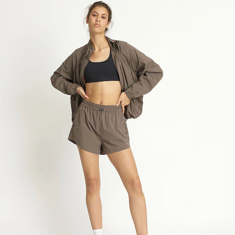 Eine Frau posiert vor einem schlichten Hintergrund und trägt eine braune Jacke, passende braune Shorts, einen schwarzen Sport-BH und weiße Socken. Sie steht selbstbewusst mit einer Hand an der Hüfte, die andere hält ihre Jacke.