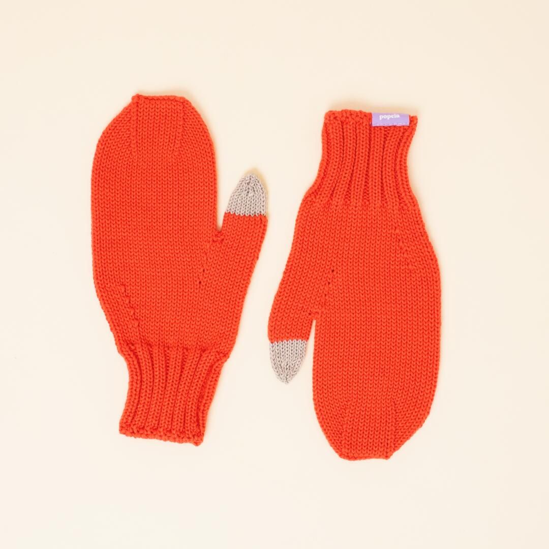 Ein Paar rote Strickfäustlinge mit gerippten Bündchen, jeweils mit einer grauen Fingerspitze am Daumen, flach auf einem hellbeigen Hintergrund.