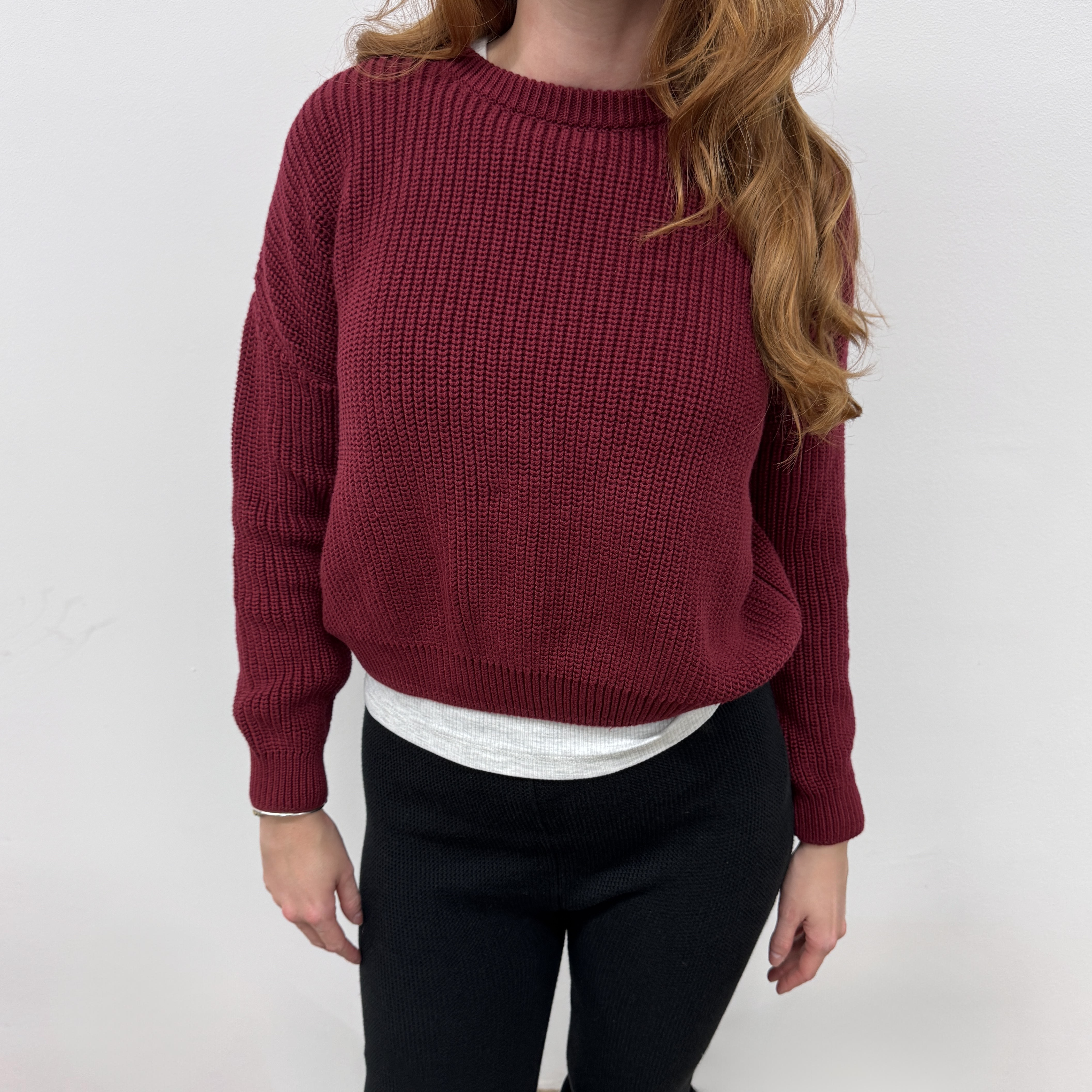 Eine Frau mit langem, gewelltem, kastanienbraunem Haar trägt einen kastanienbraunen Strickpullover über einem weißen Oberteil und eine schwarze Hose und steht vor einem schlichten weißen Hintergrund. Ihr Gesicht ist nicht zu erkennen.