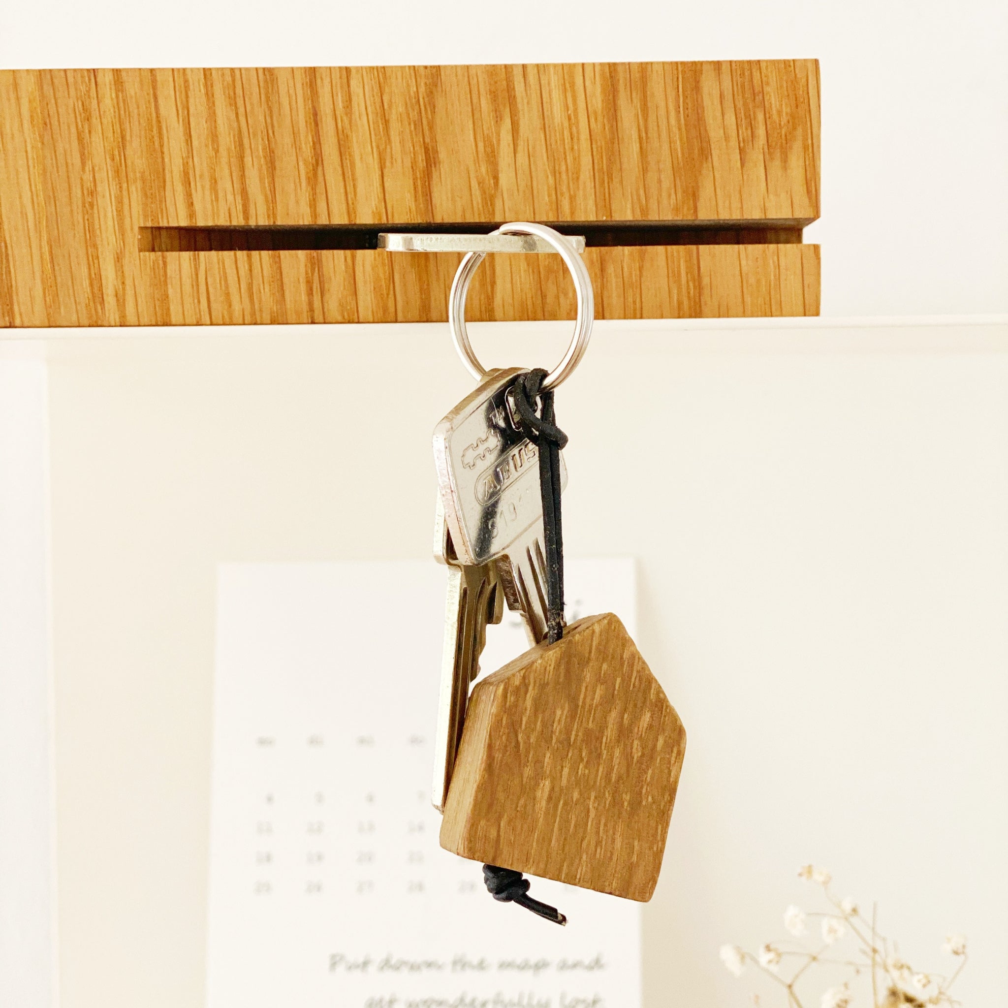 Ein Schlüsselbund mit einem hölzernen hausförmigen Schlüsselanhänger, der an einem hölzernen Haken hängt, mit einem verschwommenen Kalender und einigen weißen Blumen im Hintergrund.
