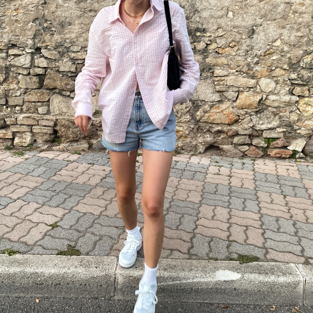 Eine Person geht auf einem gemusterten Steinweg, trägt ein rosa kariertes Hemd, Jeansshorts, weiße Socken und weiße Turnschuhe und hat eine schwarze Tasche über der Schulter. Im Hintergrund ist eine Steinmauer zu sehen.