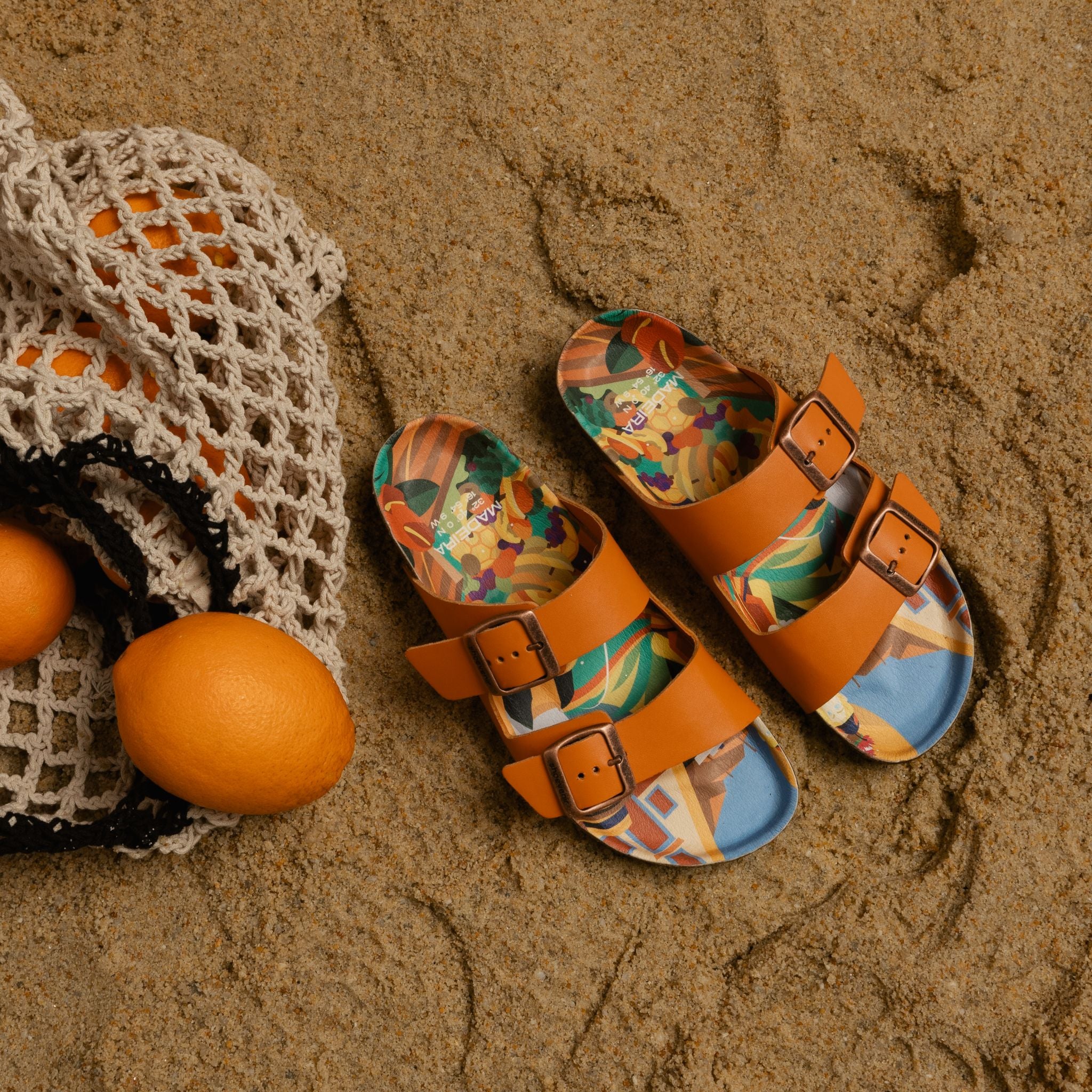Ein Paar hellbraune Sandalen mit bunten Cartoon-Sohlen liegt auf dem sandigen Boden neben einer weißen Netztasche mit zwei Orangen.