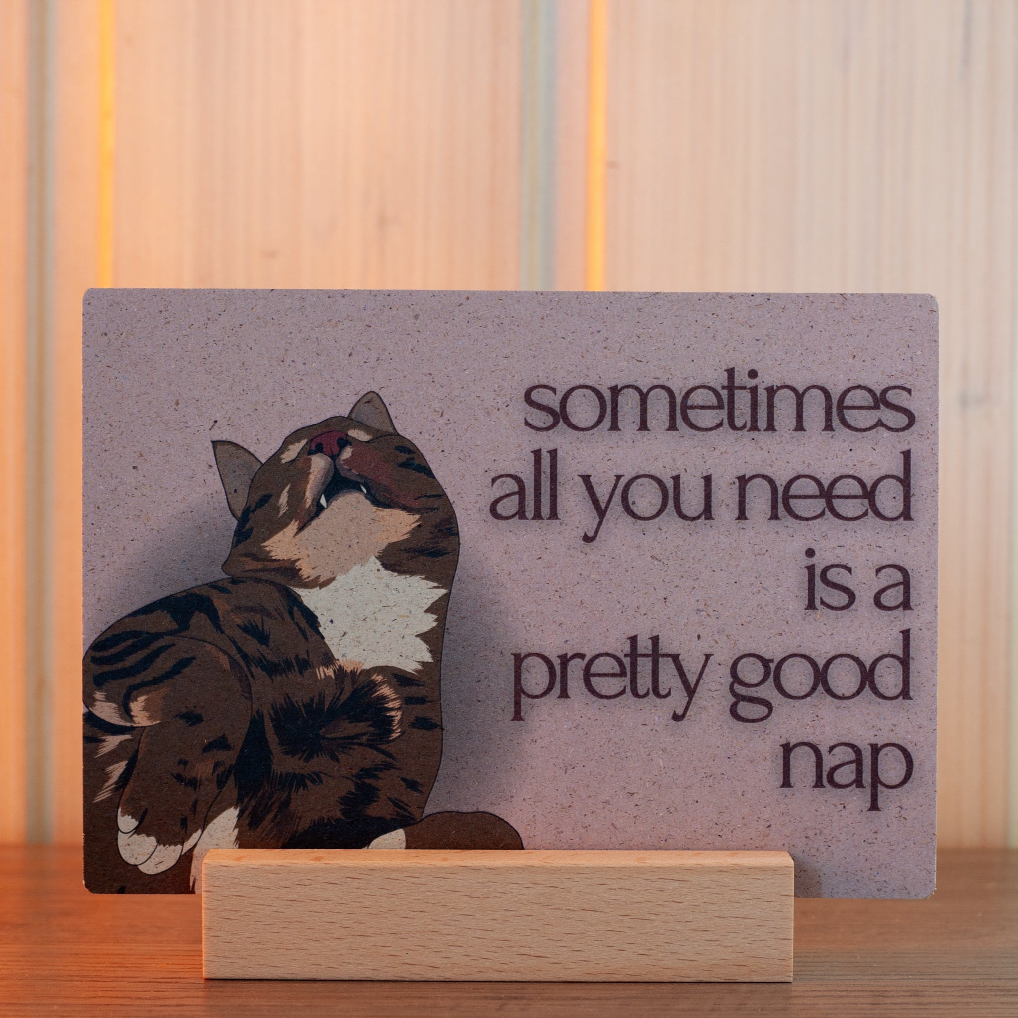 Eine Karte mit der Illustration einer entspannten, braun getigerten Katze und dem Text "manchmal ist alles, was man braucht, ein gutes Nickerchen", die auf einem Holzständer vor einem hellen Holzhintergrund präsentiert wird.