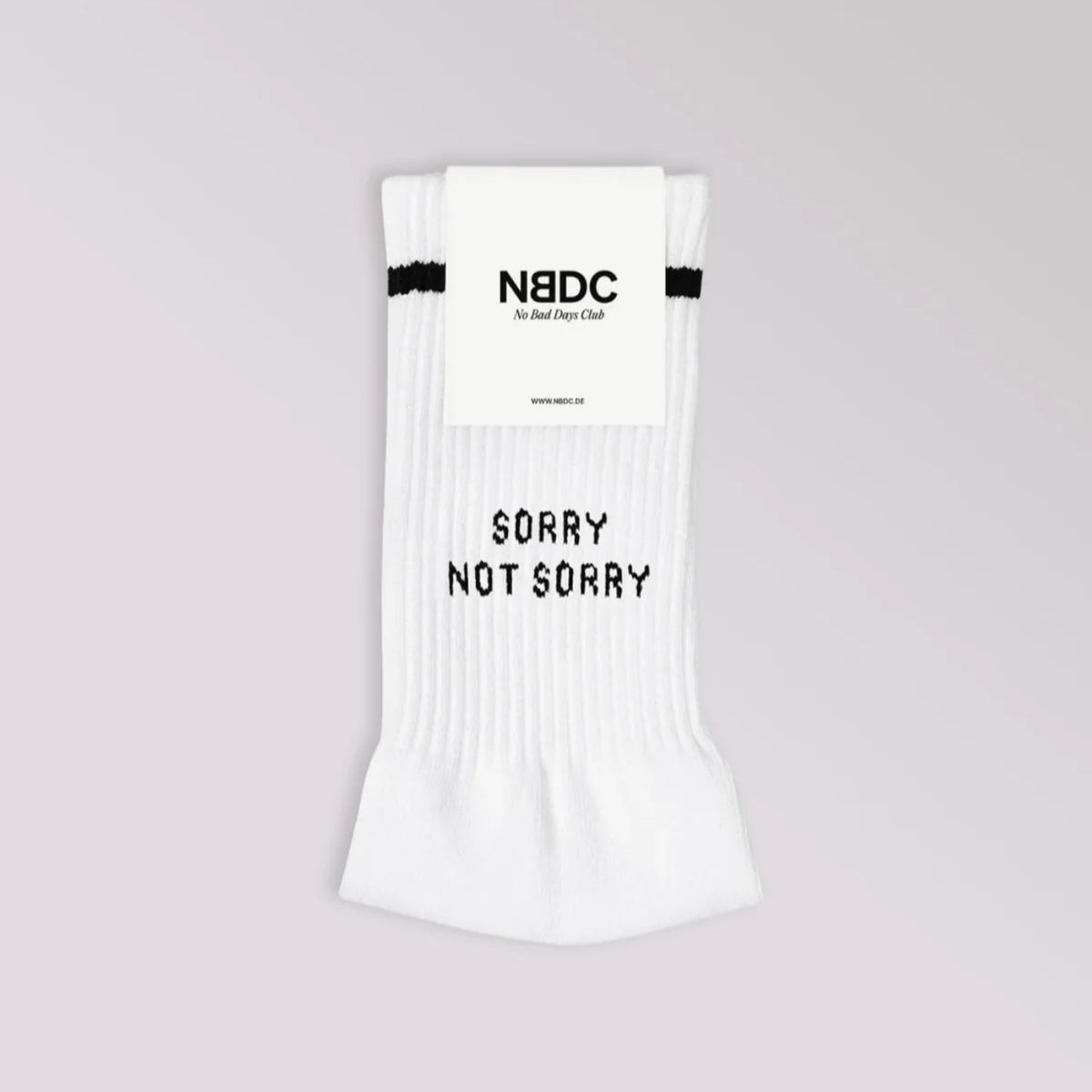 Ein Paar weiße Socken mit einem schwarzen Streifen am oberen Ende und der Aufschrift SORRY NOT SORRY. Die Socken sind gefaltet und tragen oben ein Etikett mit der Aufschrift NBDC - No Bad Days Club. Der Hintergrund ist hellgrau.