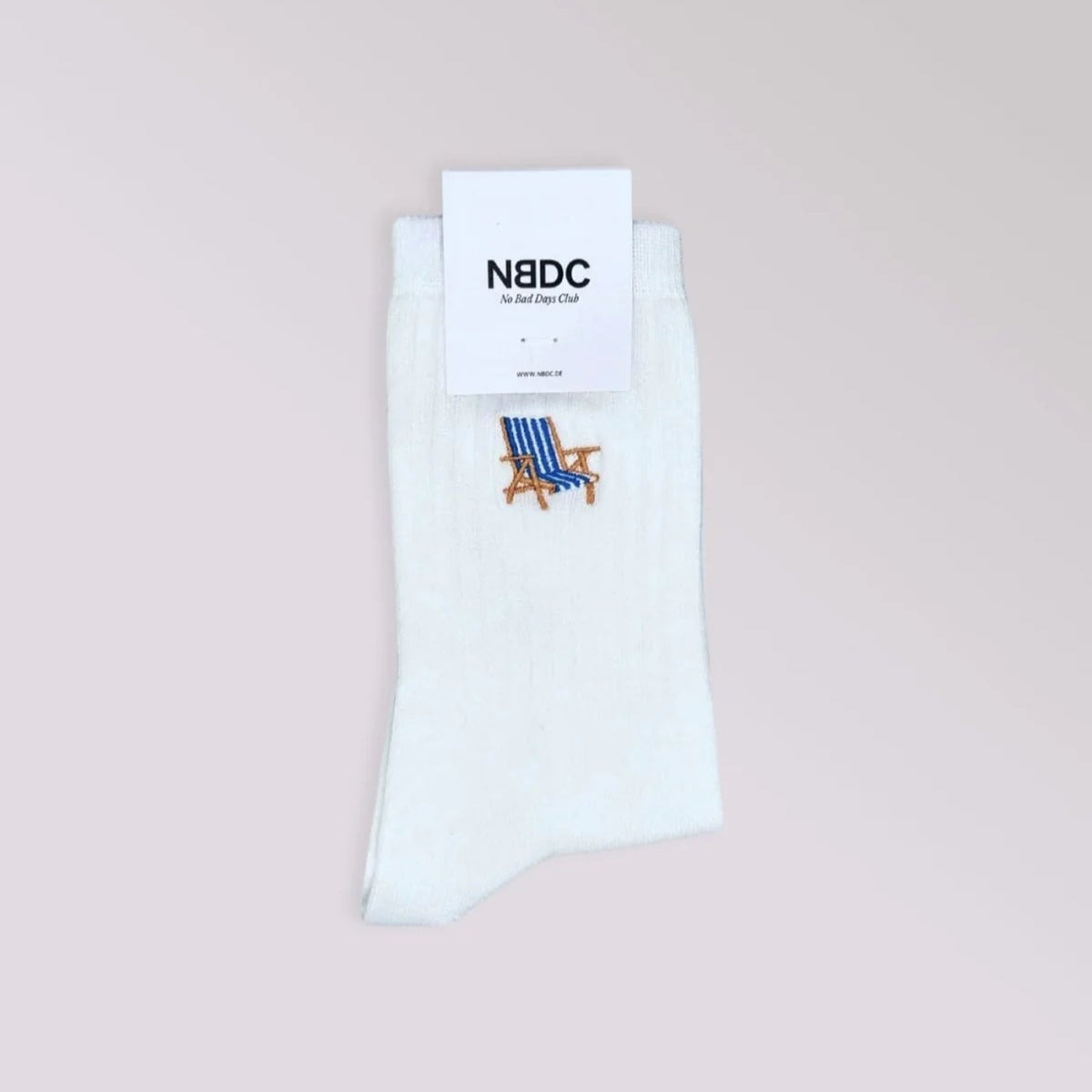 Ein Paar weiße Socken mit einem kleinen gestickten blauen und braunen Strandkorbmuster auf hellgrauem Hintergrund. Ein Etikett am oberen Rand lautet "NBDC".