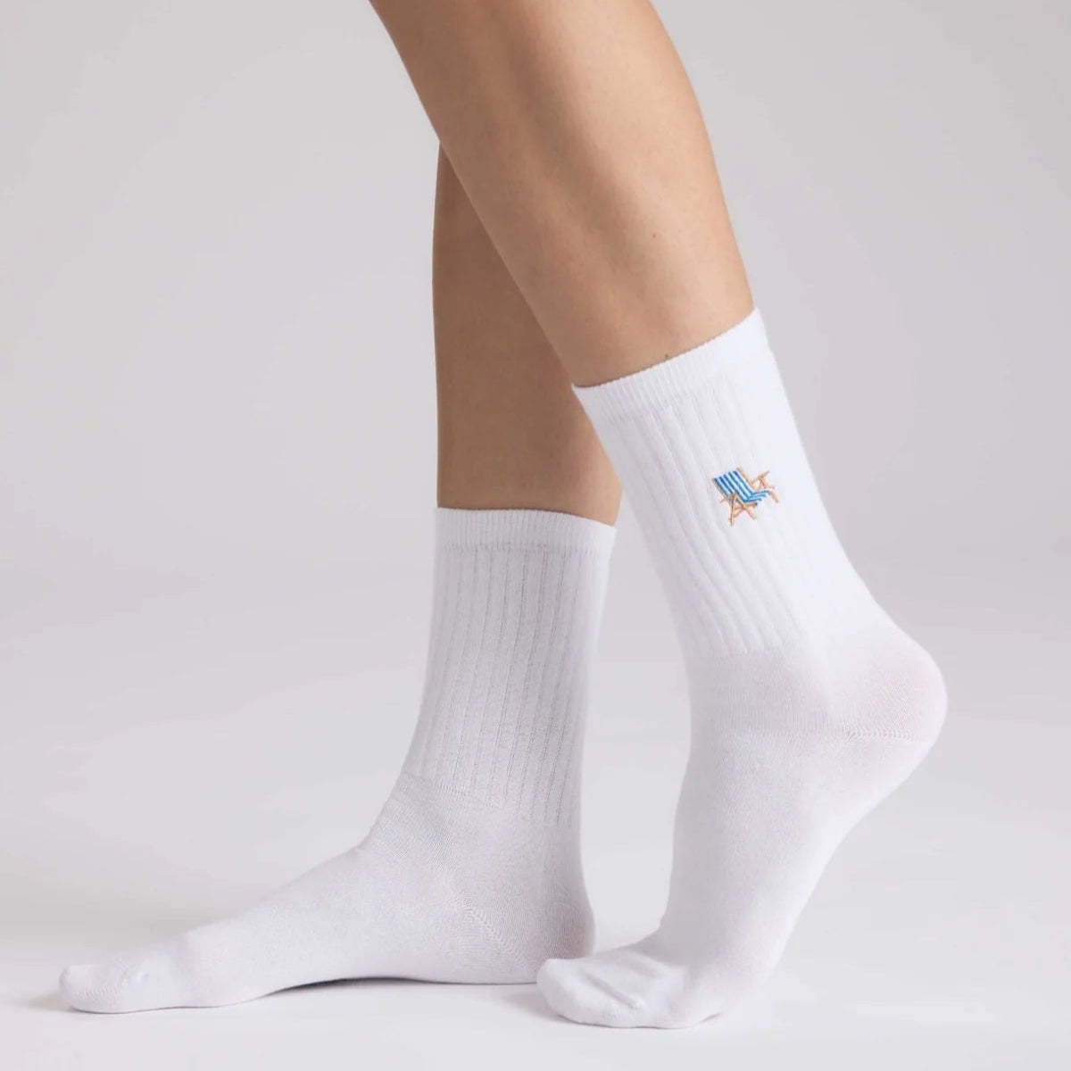 Eine Person, die weiße Socken mit einem kleinen bunten gestickten Dinosaurier an der Seite trägt, steht auf einem weißen Hintergrund.