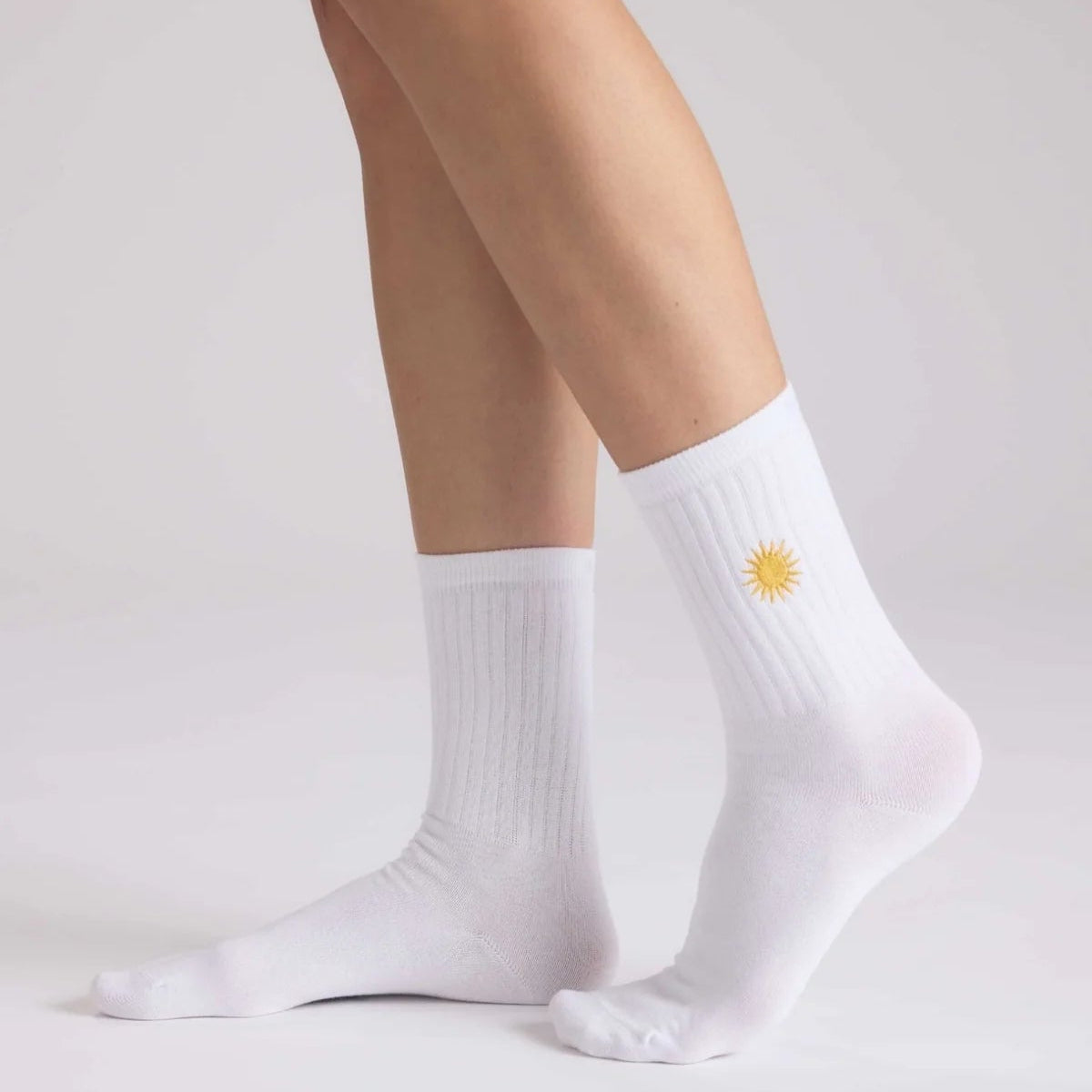 Eine Person, die weiße gerippte Socken mit einem kleinen gelben Sonnenmuster am oberen Rand trägt, steht auf einer hellen Fläche.