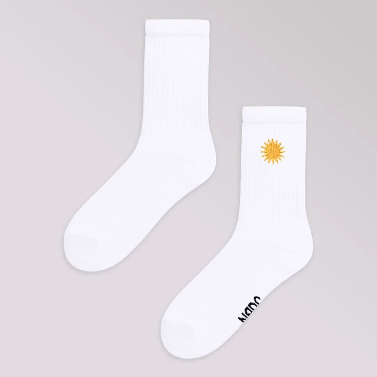 Ein Paar weißer Socken ist auf einem hellgrauen Hintergrund abgebildet; eine Socke weist ein kleines gelbes Sonnenmotiv im oberen Bereich auf und die andere hat einen schwarzen Text im Zehenbereich.