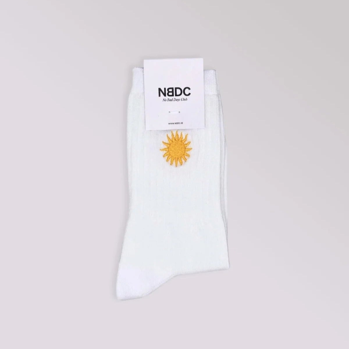 Ein Paar weißer Socken mit einem gelben Sonnenmotiv ist vor einem hellgrauen Hintergrund abgebildet. Die Socken haben oben ein Etikett mit der Aufschrift NBDC.