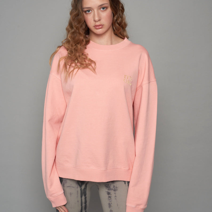 Junge Frau mit lockigem Haar trägt einen rosa Pullover und gemusterte Jeans vor grauem Hintergrund.