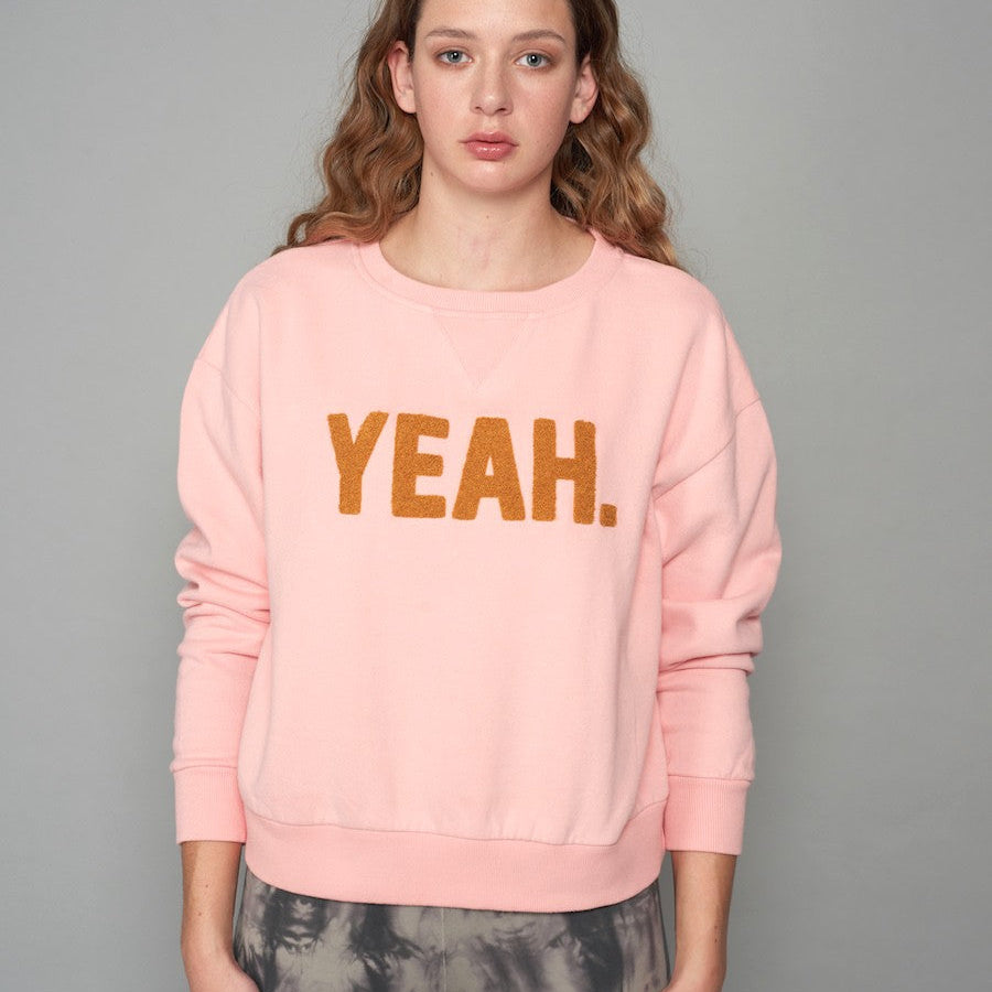 Person in a pink sweatshirt with "YEAH." written in brown letters, standing against a gray background.