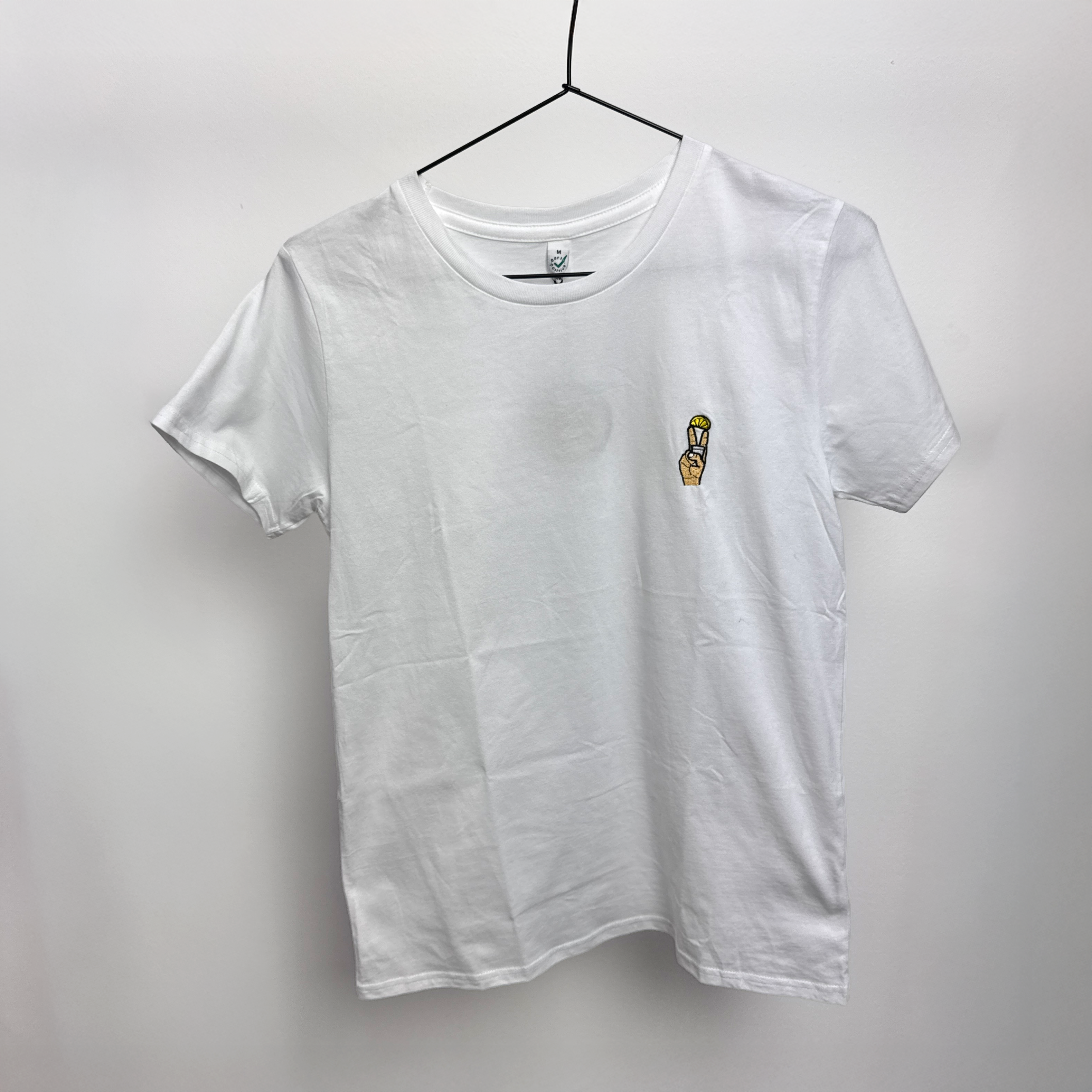 Ein schlichtes weißes T-Shirt hängt auf einem schwarzen Bügel vor einem weißen Hintergrund. Auf der linken Brust befindet sich eine kleine gestickte Grafik einer Person in Gelb, die ein Friedenszeichen hochhält.