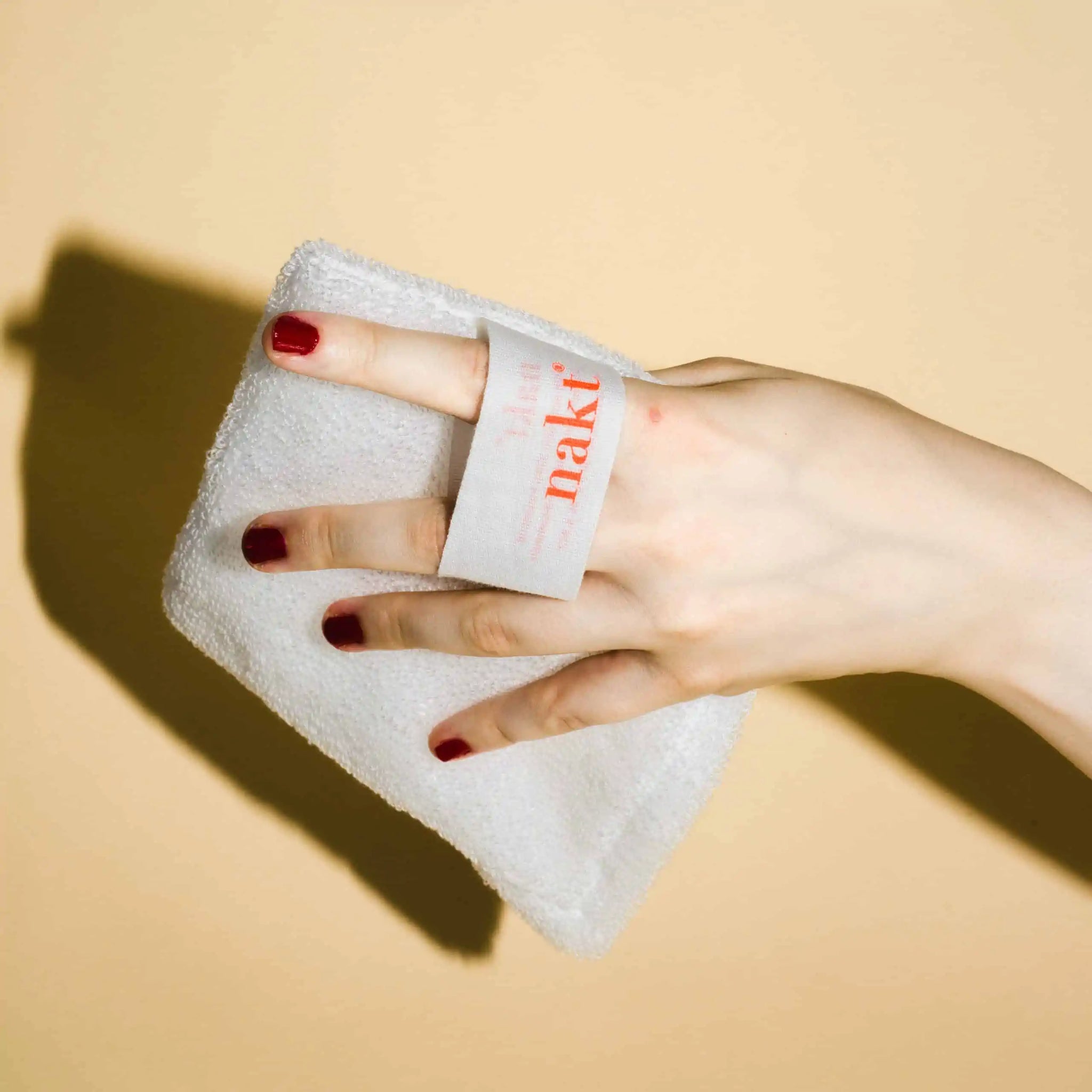 Hand mit rot lackierten Nägeln hält weißen Waschlappen mit der Aufschrift "naki" auf beigefarbenem Hintergrund.